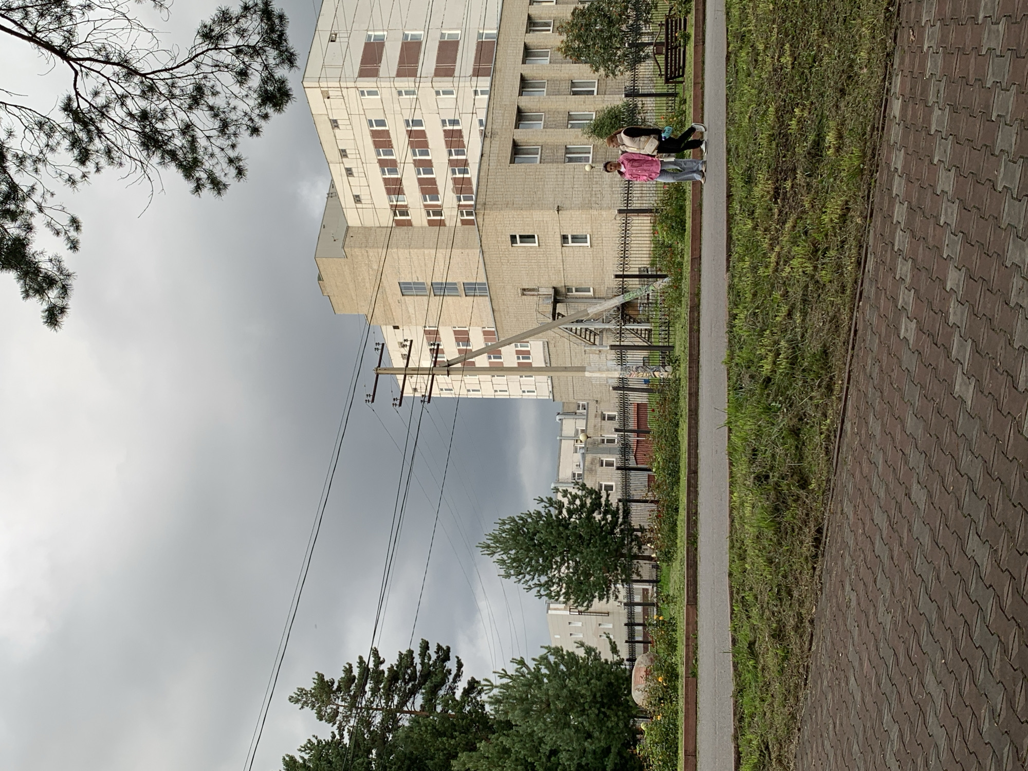 The image shows a view of buildings and trees under a cloudy sky. Two individuals, appearing to be women, are walking along a paved path in front of a black metal fence. One woman is wearing a pink jacket and blue jeans, and the other is wearing a beige vest over a light-colored top and dark pants. Beyond the fence, there are several trees, some with green foliage, and some buildings that appear to be institutional, possibly part of a hospital or a school complex in Prokopyevsk, Russia. The buildings are multi-story, with a mix of brick and lighter-colored facades, and numerous windows. There are power lines visible crisscrossing the sky. In the foreground, there is a paved area with red and gray interlocking bricks, and a strip of mowed grass. The overall atmosphere is overcast and slightly somber.
