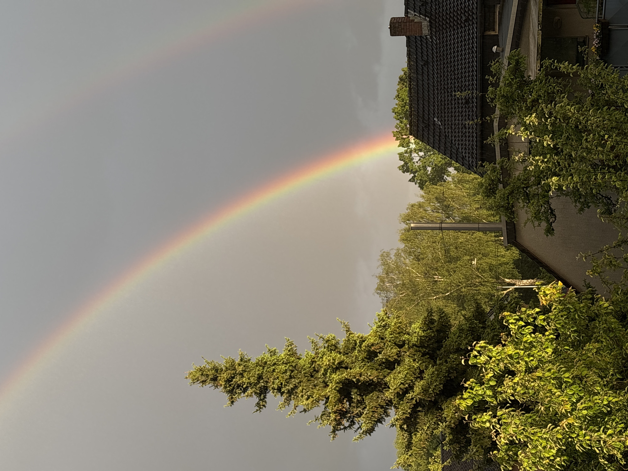 A rainbow arches across the sky above a neighborhood in Bochum, Germany. The rainbow's vibrant colors contrast with the overcast sky. The base of the rainbow appears to descend into a lush green area, bordered by trees and a house with a dark tiled roof and chimney. A secondary, fainter rainbow is visible above the main one. A street lamp is seen amidst the trees.