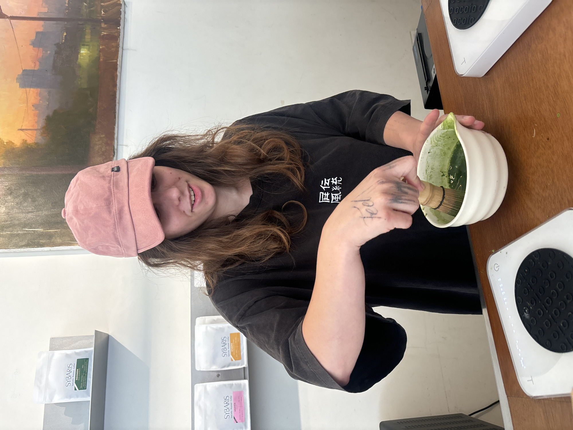An indoor, vertical-format photograph captures a person preparing matcha, likely in a cafe or specialty beverage establishment in Tel-Aviv, Israel. The subject, a young individual with long brown hair, is wearing a pink baseball cap and a black t-shirt. They are smiling faintly while using a bamboo whisk to froth green matcha in a white, ribbed bowl. A tattoo reading "Feel" is visible on their right forearm, and the black t-shirt features white text that appears to be "屏伝" and "風統" (likely Chinese or Japanese characters).
The person is positioned behind a wooden counter, which holds an Acaia digital scale. In the background, a painting on the wall depicts a cityscape with warm, orange hues, suggesting either sunrise or sunset. To the left, several white pouches of coffee beans are displayed, clearly labeled "SYBARIS Specialty Roasted Coffee." Specific origins like "INDONESIA" and "UGANDA" are discernible on the bags. The setting suggests a focus on artisanal coffee and tea.