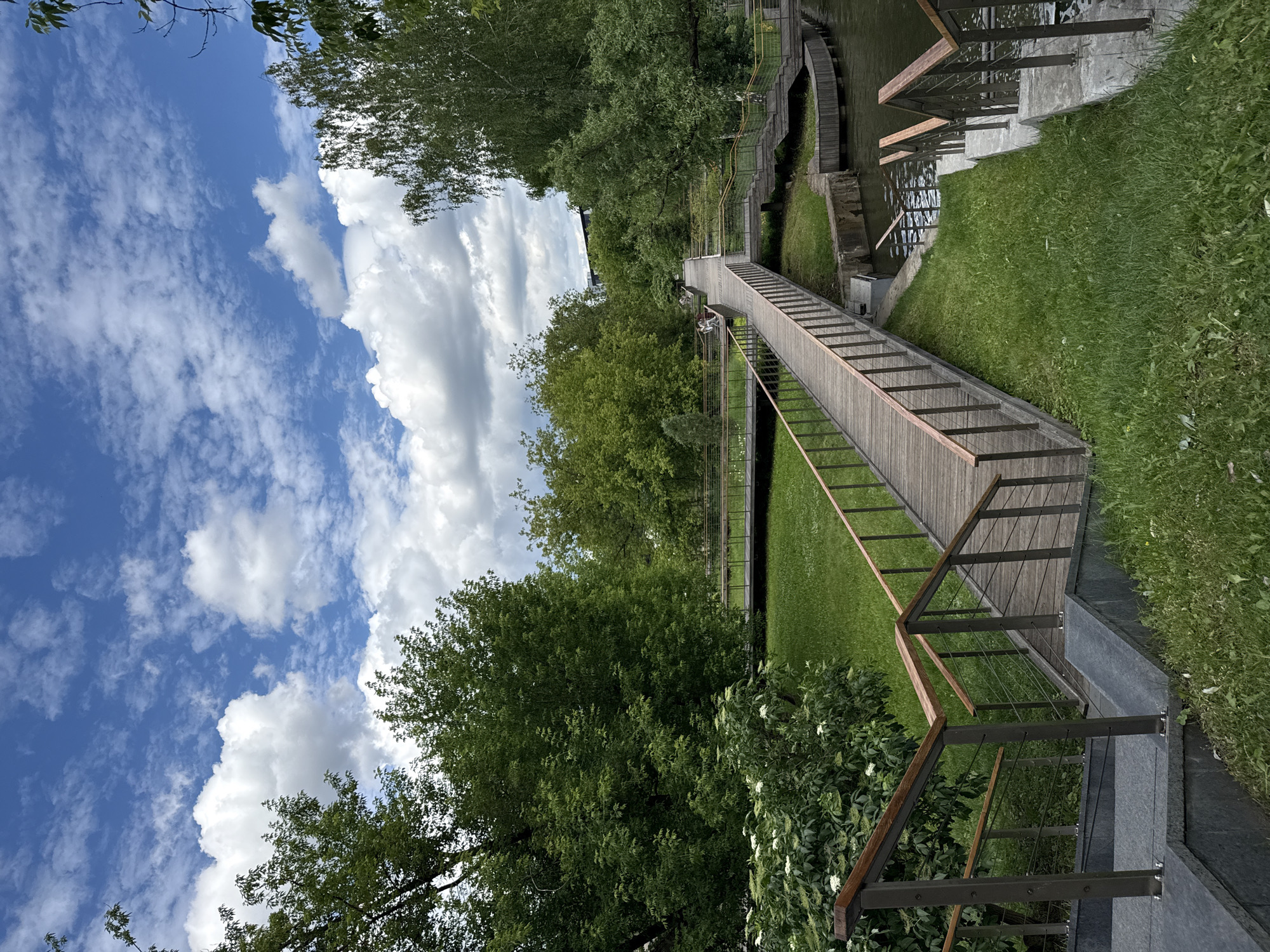 The image depicts a park with a wooden footbridge over a body of water. The bridge has railings and stairs leading down to the water's edge. The area is surrounded by lush green grass and trees. The sky is blue with scattered white clouds. In the distance, there are some buildings visible. The scene appears to be from a park in Moscow, Russia. There are a few people visible on the bridge in the distance. The overall atmosphere is peaceful and serene.