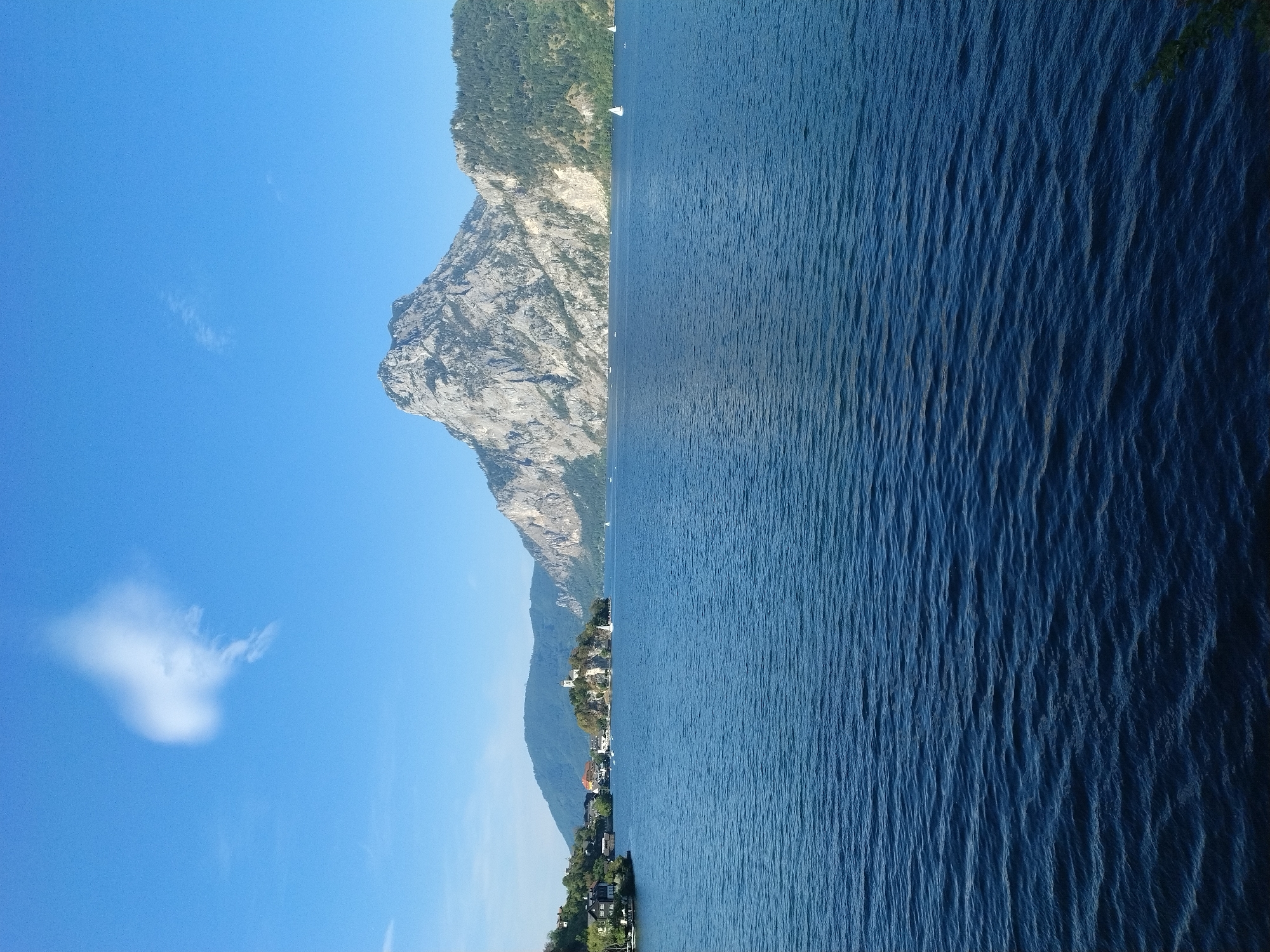 This is an outdoor, daytime, eye-level, full shot photograph of a wide, deep blue lake with gentle ripples on its surface. The lake occupies the majority of the frame, from the bottom edge to roughly the mid-point. In the background, a prominent, jagged, grey mountain with sparse green trees on its slopes dominates the right side of the image. To the left of the mountain, a cluster of green, tree-covered hills slopes down towards the water. On the shore of the lake, nestled among trees, are several buildings with red-tiled roofs and white or cream-colored walls, some of which appear to be houses. Further out on the water, a small island is visible, topped with a white building that resembles a church or castle. Several small sailboats with white sails are scattered across the lake. The sky above is a clear, bright blue with a single, wispy white cloud in the upper left quadrant. The scene depicts a picturesque natural landscape with a small settlement on the lake's edge. The location is Traunkirchen, Austria, a village situated on a peninsula in the Traunsee lake.