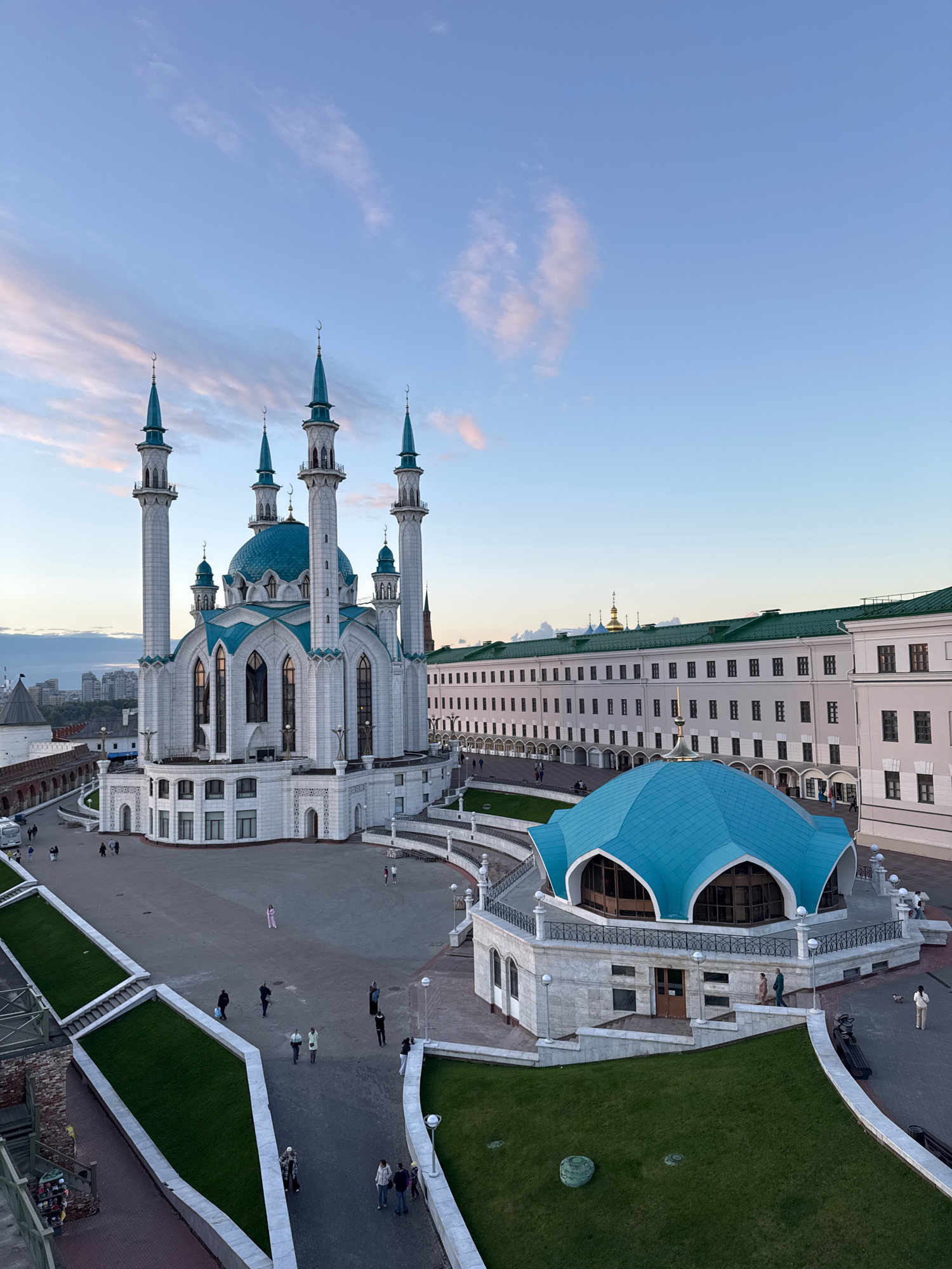 This is a high-angle, medium shot of the Kul Sharif Mosque in Kazan, Russia, taken at dusk. The sky is a gradient of pale blue and light pink, with wispy clouds. The mosque, made of white stone with blue accents, is the central subject, featuring a large dome and four tall minarets, each topped with a crescent moon. To the right of the mosque, a smaller, octagonal building with a bright blue, petal-shaped roof is visible. In the background, a long, pale building with a green roof and numerous windows stretches across the frame. The foreground shows paved courtyards and grassy terraces. Several individuals are scattered throughout the scene, walking on the pathways and plazas. One person in a pink outfit is standing in the middle of the main plaza. Another person is sitting on a bench near the blue-roofed building. The lighting suggests the sun has recently set, casting soft shadows.