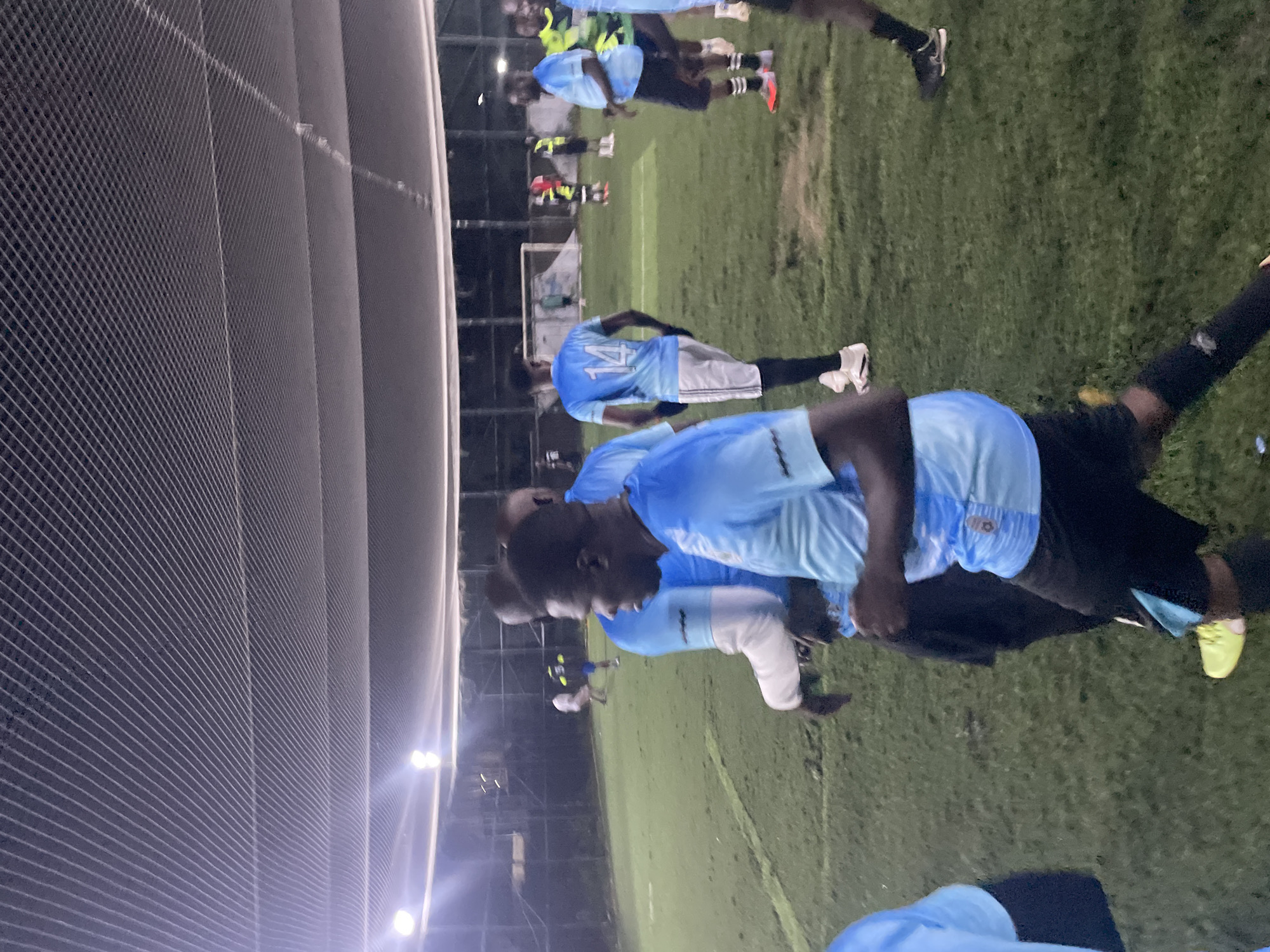 The image shows a group of men in light blue soccer jerseys on a turf field at night, likely in Gura Top, Nigeria. The field is enclosed by a net ceiling and illuminated by artificial lights. Several men are visible in the foreground and midground, some walking and others standing. One man in the foreground is wearing a light blue jersey with white trim and black shorts, with the number 14 visible on his back. Other players in the background are also wearing light blue jerseys. The overall scene suggests a casual soccer game or practice taking place in an urban environment.