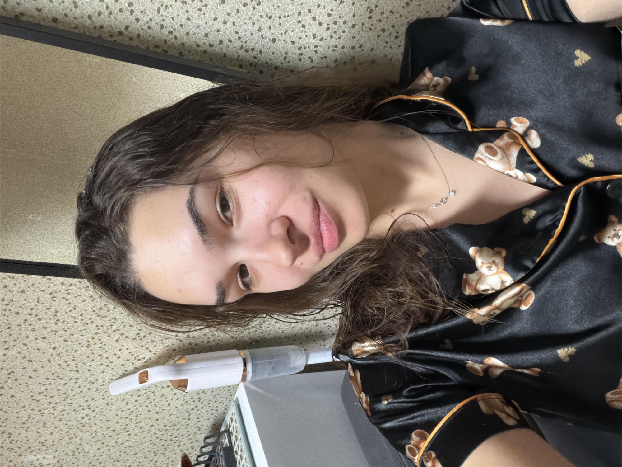 A young woman with damp, dark brown hair lies sideways on a textured surface, likely a bed or couch. She is wearing a black silky pajama top with a teddy bear print and orange trim, and a delicate silver necklace with a small pendant is visible. The woman's face is visible and she is looking into the camera. Next to her is a white nebulizer or vaporizer device. The exact location in Sofia, Bulgaria, cannot be determined from the image.