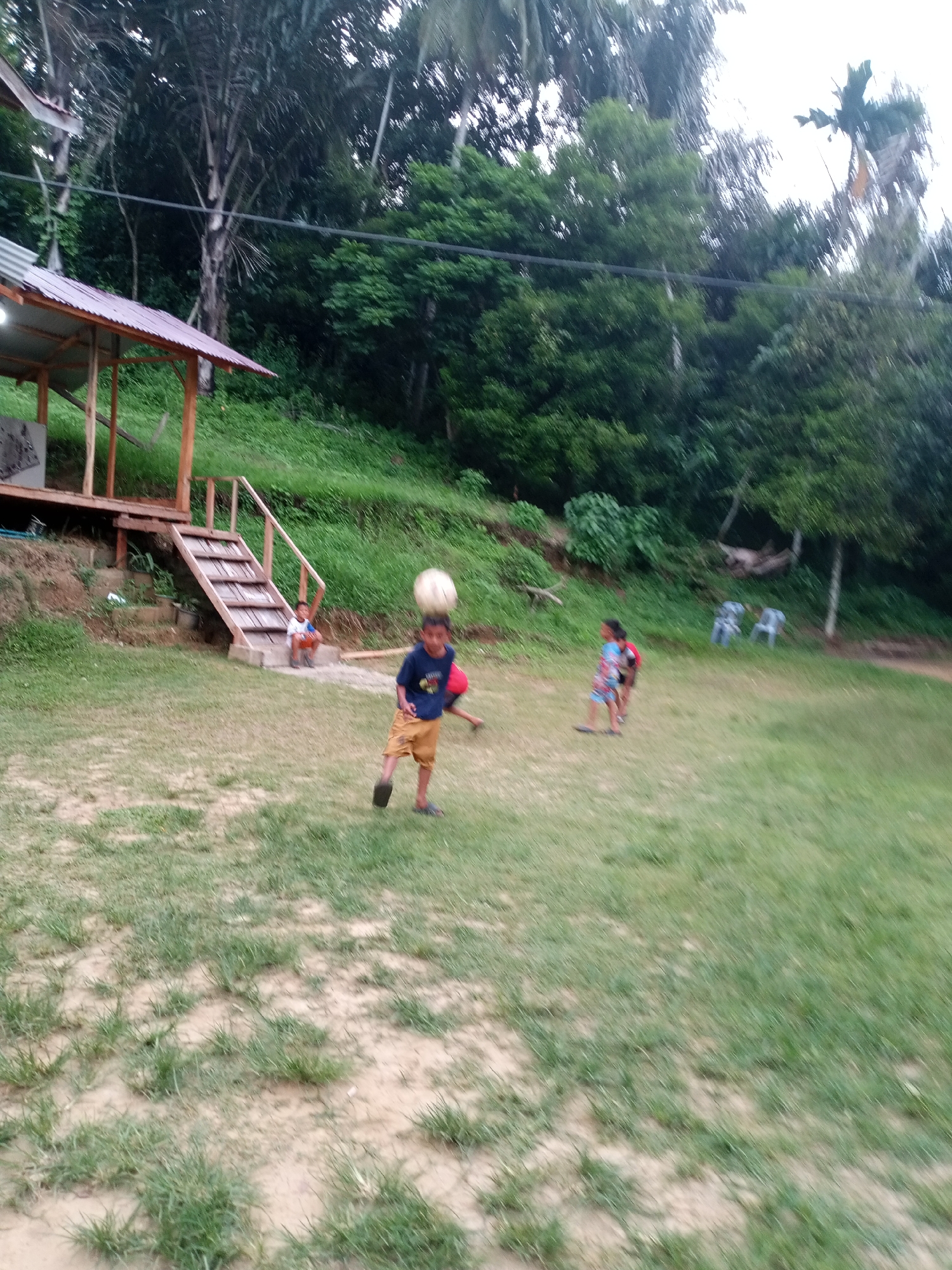 This media file depicts a group of children playing on a grassy field near a wooden structure. The main subject in the foreground is a young boy wearing a dark blue t-shirt and yellow shorts, actively engaged in playing with a soccer ball that is hovering in the air above his head. He appears to be in motion, perhaps mid-kick or receiving the ball. To his left, further back on a set of wooden stairs leading up to a covered structure, sits another young boy wearing a white t-shirt and orange shorts. In the middle ground, two more children are visible. One, to the right of the main subject, is wearing a red and blue patterned outfit and appears to be running or kicking a ball. Another child, further in the background, is partially obscured but seems to be wearing a red shirt and blue shorts.
The setting is an outdoor environment with a focus on a well-maintained grassy area. Behind the children, a lush green hillside rises, covered in dense foliage and trees, suggesting a tropical or sub-tropical location. A simple wooden structure with a reddish-brown roof is visible to the left of the frame, with visible wooden beams and a staircase. In the background, several white plastic chairs are scattered on the grass. The lighting suggests it is daytime, likely late afternoon given the soft light and the overall ambiance. The overall mood conveyed is one of joyful activity and outdoor play. There is no visible text in the media file. The location context is Lhokseumawe, Indonesia.
