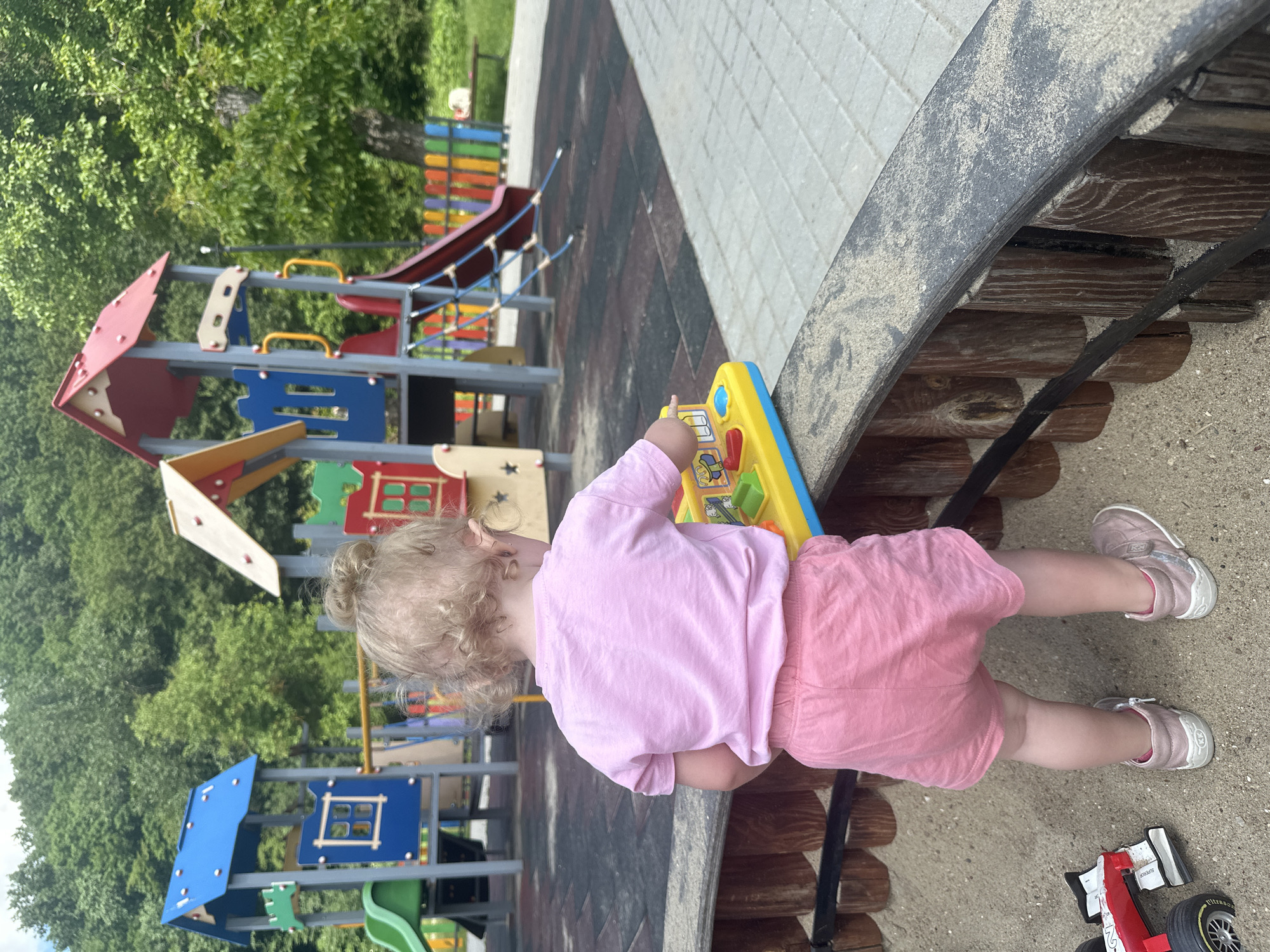 A blonde toddler with curly hair, seen from behind, is playing in a sandbox at a playground in Donchovci, Bulgaria. The child is wearing a pink t-shirt and pink shorts, and pink shoes. In front of the child is a yellow toy with colorful buttons and levers. A red and white toy car is visible to the left in the sandbox. The playground features a colorful slide, climbing structures, and a fence. The ground is a mix of rubber matting and paved tiles. The background consists of lush green trees.