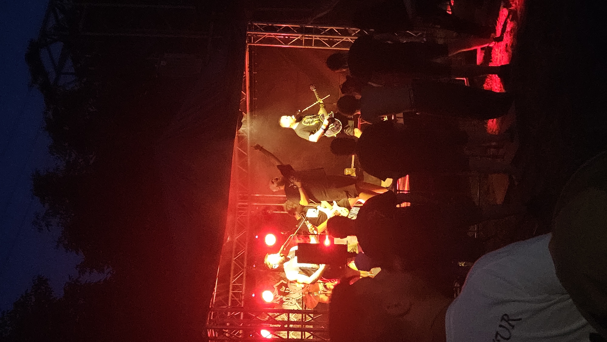 The image depicts a live outdoor music performance at night, illuminated by red stage lighting. A band of at least four members is performing on a raised stage constructed with metal scaffolding and draped with a dark backdrop. The band members are dressed in casual, dark clothing. One vocalist is at the front center, singing into a microphone. To his right, a guitarist is playing an electric guitar. To the left of the vocalist, another band member is partially visible, possibly playing drums or another instrument. The stage is set up in an open area with trees visible in the background against a dark sky. In the foreground, silhouetted figures of audience members are visible, facing the stage. The ground around the stage is dimly lit by the red stage lights, casting a red glow on the grass. The overall atmosphere suggests a rock or punk music concert. The location is not explicitly stated in the image.