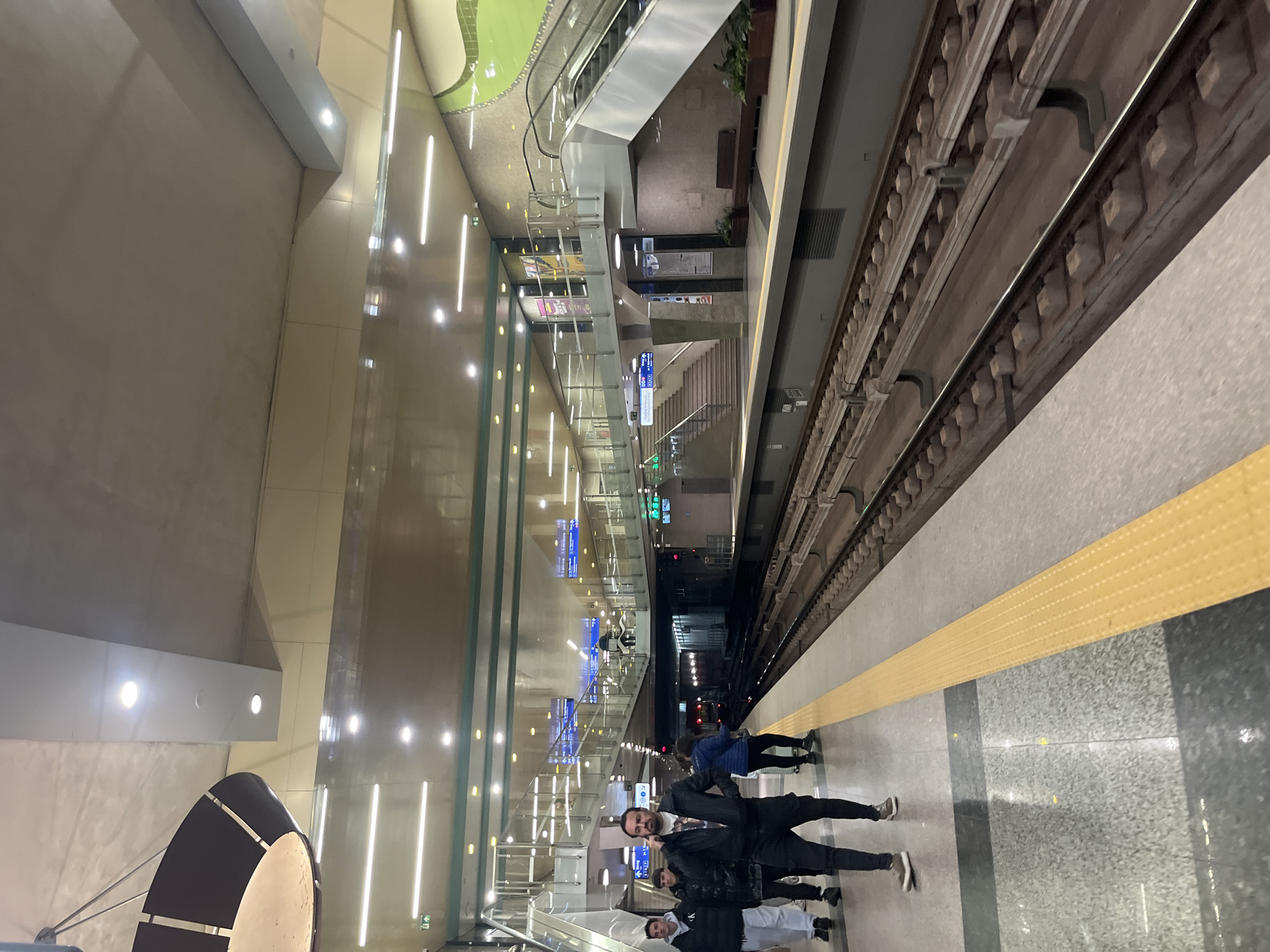 This image captures a modern, brightly lit underground metro station platform, likely in Sofia, Bulgaria, given the context. The perspective is from the platform level, looking across the tracks towards the opposing platform and up towards a multi-level entrance/exit area.
The station features clean, contemporary architecture with smooth, light-colored walls and ceilings. A distinctive light green, undulating decorative element is visible on an upper level, complementing the overall bright aesthetic. The station is illuminated by numerous rectangular and circular LED lights embedded in the ceiling and walls.
On the right side of the image, train tracks are visible, laid on a concrete bed with exposed sleepers. A yellow tactile warning strip runs along the edge of the platform, separating the waiting area from the tracks.
In the foreground on the platform, several people are standing, appearing to wait for a train. A group of four individuals is prominent, with one man in a black jacket and a beard looking directly towards the camera. Another person in a blue jacket stands further down the platform.
To the left and back, the station's upper levels are accessible via escalators and stairs, framed by glass barriers. Blue information signs with white text and symbols are mounted throughout these areas, likely providing directions or station information. Some potted plants are visible near the escalators, adding a touch of greenery. In the bottom left, a large, dark, curved light fixture or decorative element is partially visible. The overall impression is one of a clean, efficient, and well-maintained public transport space.