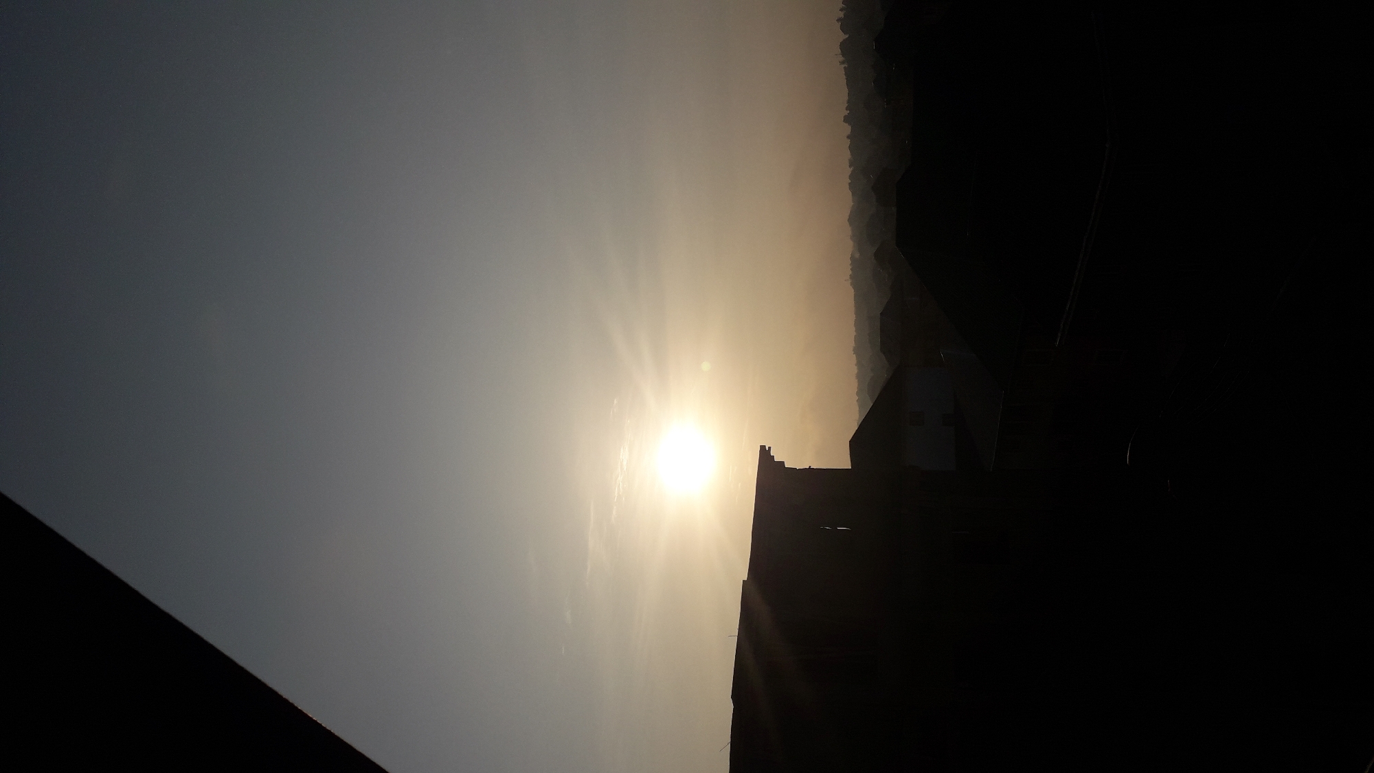 The image captures a sunset scene in Nkpor, Nigeria. The sun, a bright orb, is positioned in the upper-middle section of the frame, casting a warm glow across the sky. Its rays extend outwards, creating a starburst effect. In the foreground, the silhouette of a building with a flat roof dominates the left side of the image, its dark form contrasting sharply with the lighter sky. Beyond this, a hazy silhouette of residential buildings with pitched roofs stretches across the middle ground, suggesting a residential area within the city. The overall atmosphere is serene, with the dimming light creating a peaceful ambiance. The sky transitions from a pale yellow near the horizon to a muted grey at the top, with subtle cloud formations visible. The foreground building appears to be a multi-story structure, possibly an apartment block or commercial building, casting shadows over the lower portion of the frame. The perspective suggests the photo was taken from a higher vantage point, looking out over the city.