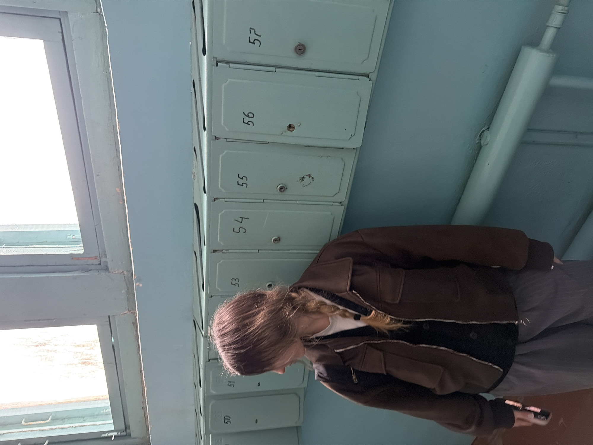 An indoor, vertically oriented shot captures a person standing in what appears to be a residential building hallway. On the right, a series of light blue-green metal mailboxes are mounted on the wall, numbered sequentially from at least "50" to "57". Specifically visible numbers are "50", "51", "53", "54", "55", "56", and "57". Many of the mailboxes have small keyholes, and some show signs of wear or minor damage around the locks.
A person, with long, light brown hair styled with a braid, is positioned in the lower center, looking down. They are wearing a brown zip-up jacket over a white top and a dark undershirt, paired with grey pants. Their left hand holds a dark-colored smartphone.
To the left, a large window allows bright daylight to flood the space, indicating it is daytime. The window frame is painted the same light blue-green as the wall and mailboxes. A white vertical pipe runs along the wall on the far right. The overall aesthetic suggests a utilitarian environment common in older apartment buildings, consistent with the provided location context of Noviy, Russia.