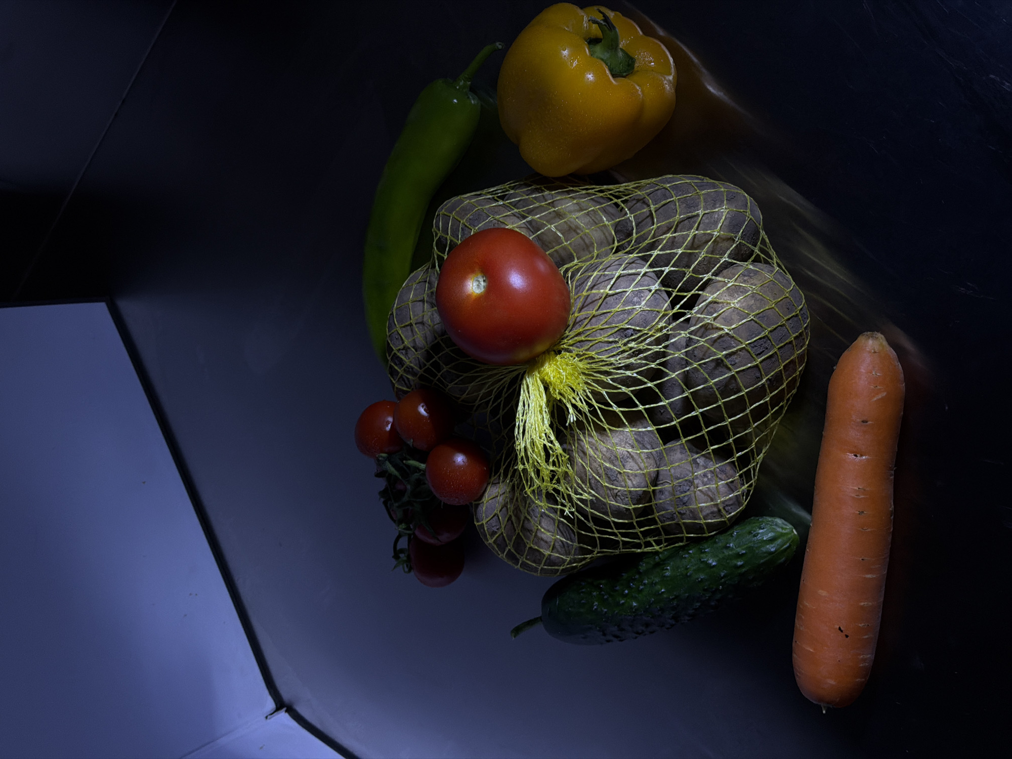 The image shows a collection of vegetables on a dark, possibly metallic surface. The vegetables include a yellow bell pepper, a long green pepper, a bag of potatoes, a tomato, a bunch of cherry tomatoes, a cucumber, and a carrot. The setting and content do not provide enough information to confidently place this scene in Dilijan, Armenia. There are no people, actions or interactions visible in the image.