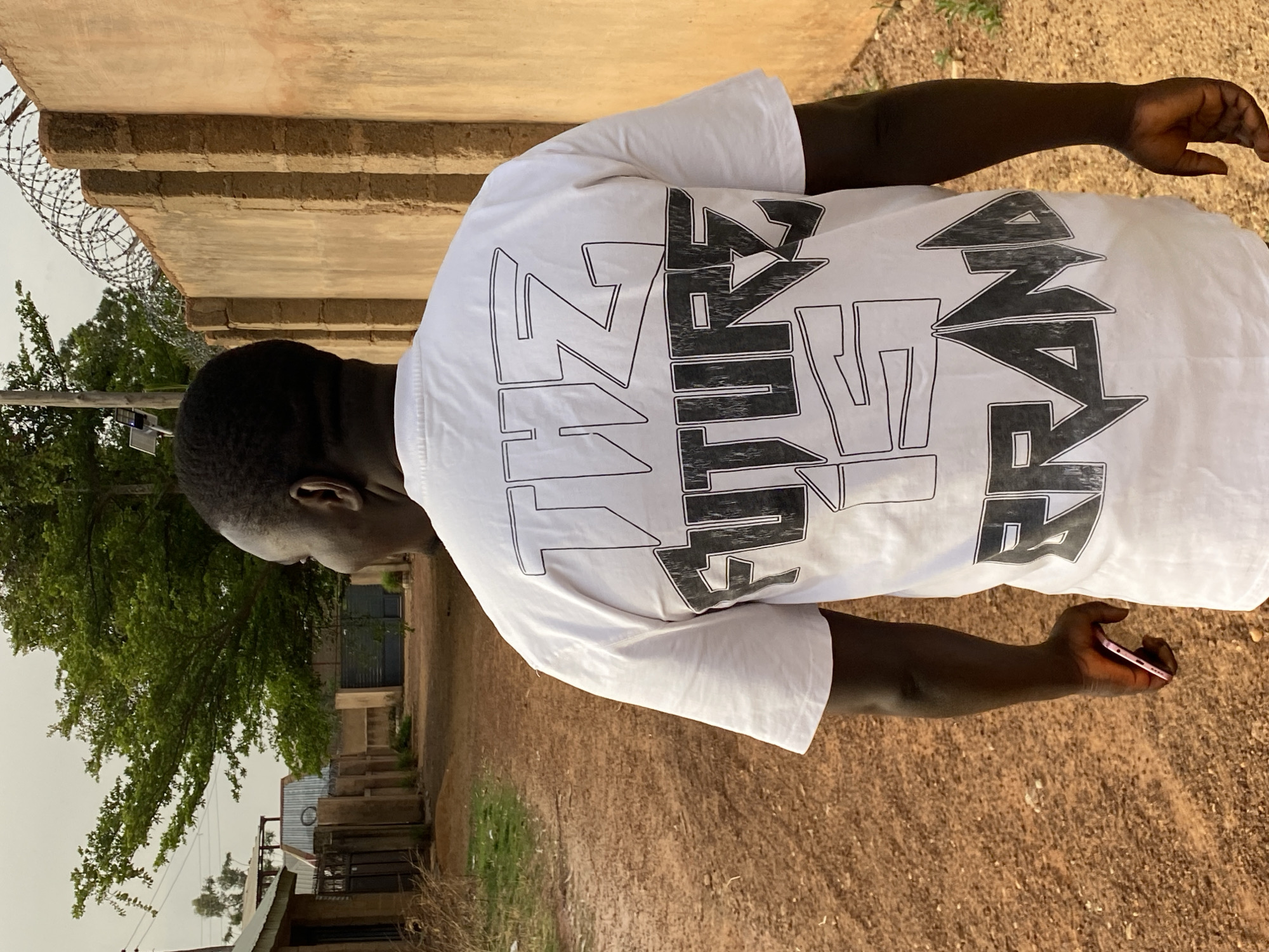 A person stands on a dirt ground in what appears to be an outdoor, possibly rural, setting. The person is wearing a white T-shirt with bold black and white text printed on the back. The text is stylized and somewhat obscured by the folds of the shirt, but "The Future Is Now" is discernible. The individual holds a cell phone in their right hand, with the screen facing away from the viewer.
Behind the person, a low wall made of brown bricks can be seen. To the left, a large tree and the corner of a building are visible. Barbed wire is situated near the wall, suggesting a perimeter or boundary.
The location is identified as Bukuru, Nigeria, although no specific landmarks or identifying features of the city are clearly visible in the frame.