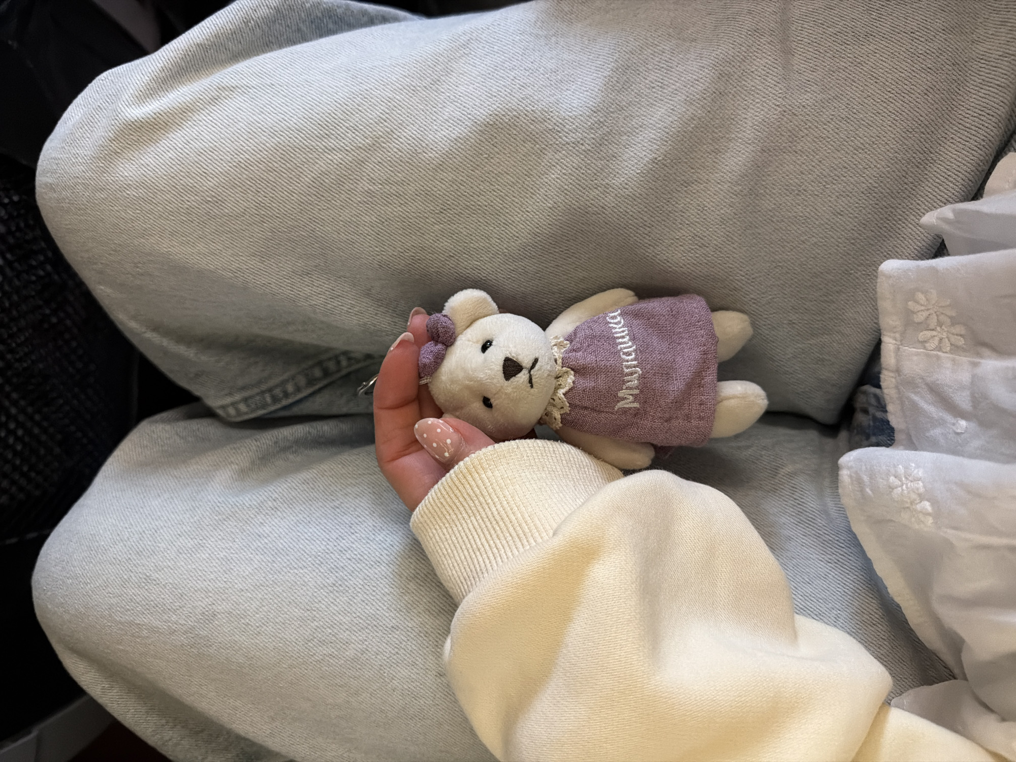A person's hand, with white nail polish dotted with light pink, holds a small white teddy bear wearing a purple dress with white embroidered text. The bear has a small purple bow on its head. The hand and bear are resting on a person's lap, clad in light blue denim jeans. The background consists of the denim jeans and the edge of a cream-colored sweatshirt or hoodie. The scene is indoors, and no specific location in Kuta, Indonesia, can be determined from the image.