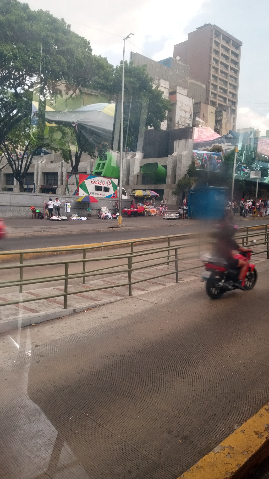 The image shows a street scene in Caracas, Venezuela, as indicated by the "Farmacia Caracas" van in the background. Several vendors have set up stalls along the street, displaying goods for sale. A person is riding a red motorcycle in the foreground. The overall scene is bustling, with many people walking around. The presence of a tall building in the background suggests a dense urban environment. The image is taken from inside a vehicle, as evidenced by reflections on the window.