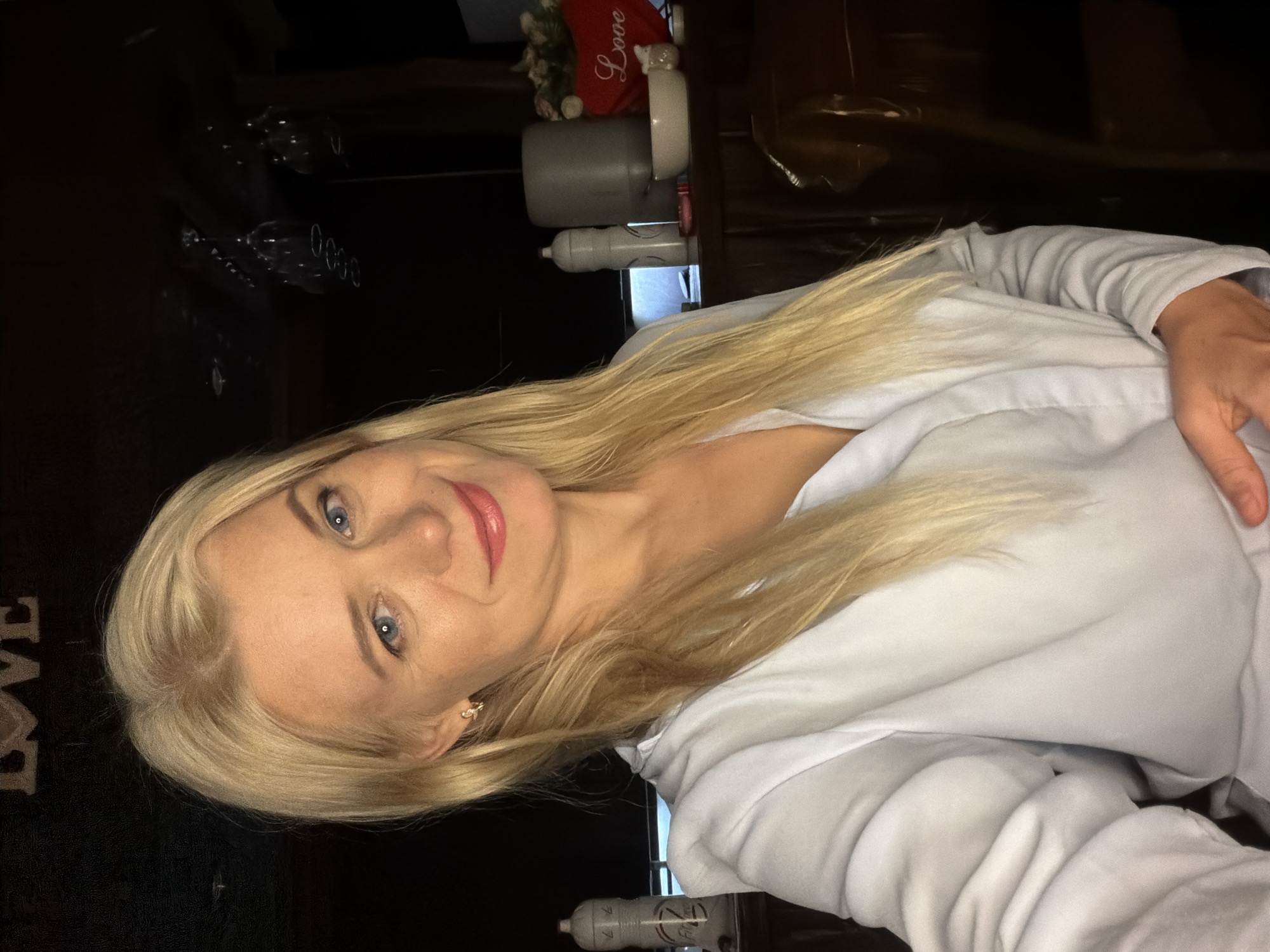 The image is a portrait of a blonde woman with blue eyes and pink lipstick. She is wearing a white hoodie and looking towards the camera with a slight smile. Her hair is long and falls over her shoulders. The background is dark, with some objects visible that are out of focus, including what appears to be a shelf with glasses and some decorative items like a red heart with "Love" written on it. A water bottle is also visible in the lower right corner of the image. The lighting suggests it might be evening or indoors. The overall mood of the image is casual and personal. The text "Love" is visible on a red decorative item. The photo appears to be taken from a slightly low angle, with the woman's head tilted.