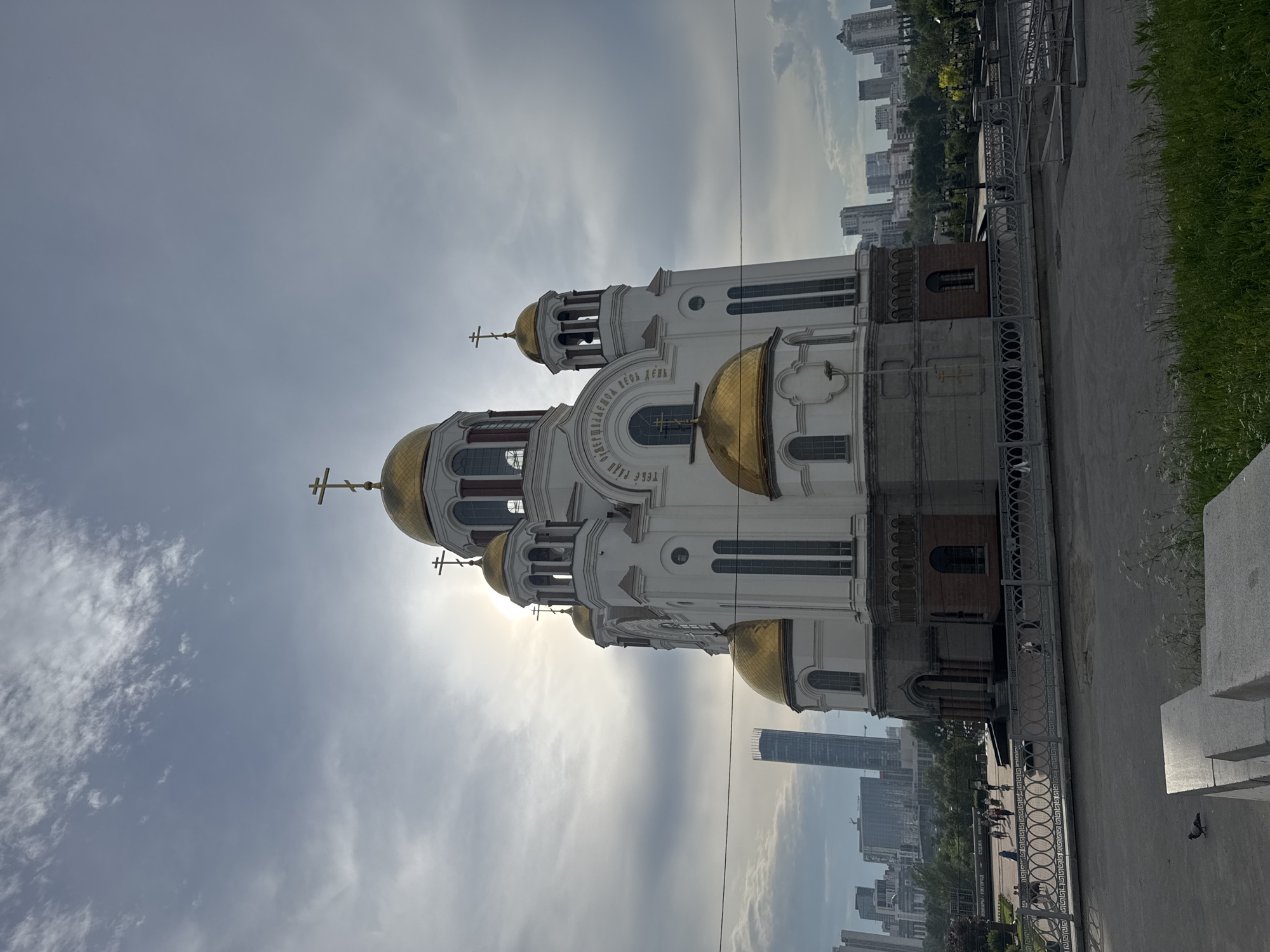 The image shows the Church on Blood in Yekaterinburg, Russia, a prominent landmark built on the site where the last Russian Emperor Nicholas II and his family were executed. The white and gold domed church stands tall against a cloudy sky, with the sun partially obscured by the clouds, creating a dramatic lighting effect. In the background, the modern cityscape of Yekaterinburg is visible, featuring skyscrapers and other buildings. A few people are seen walking in the plaza in front of the church. A pigeon is visible in the foreground on a paved area. The overall scene captures the juxtaposition of historical significance with the contemporary urban environment.