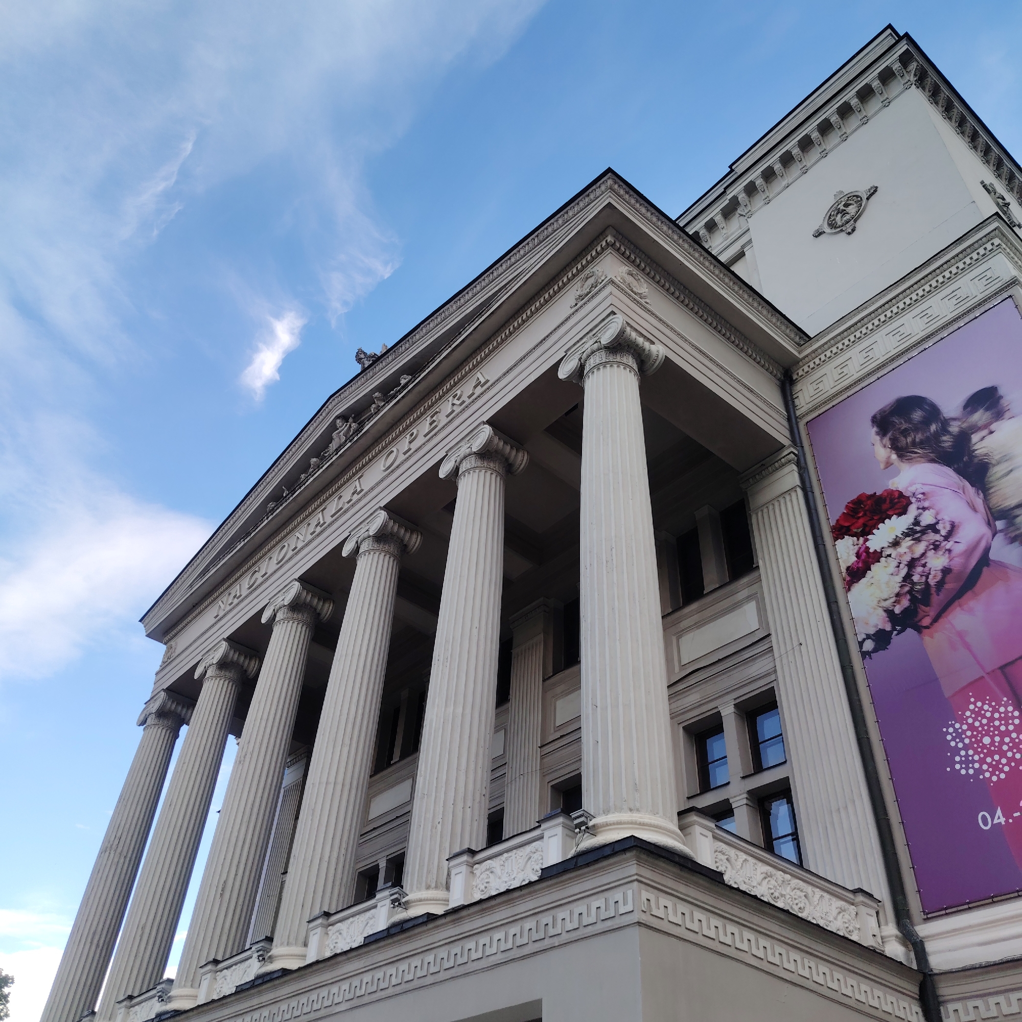 This is a low-angle, eye-level shot of the National Opera in Riga, Latvia. The building features a neoclassical facade with tall, fluted columns supporting a portico. Above the columns, carved figures are visible, and the building's name, "NACIONALA OPERA," is inscribed. A large banner depicting a woman in a pink dress holding a bouquet of flowers is attached to the right side of the building. The sky above is blue with wispy white clouds. The foreground is dominated by the architecture, with a hint of trees on the far left. The overall impression is one of grandeur and culture.