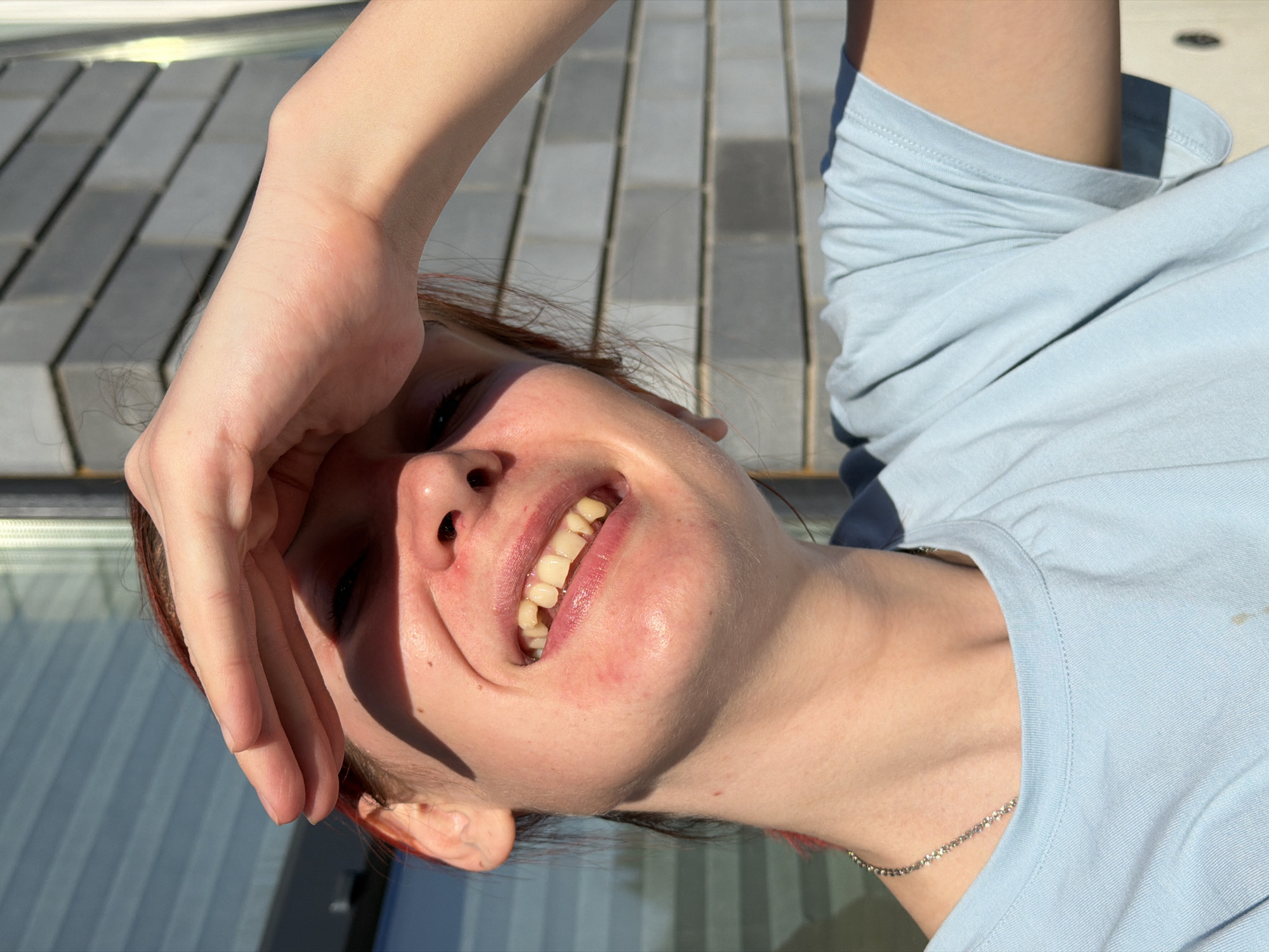 A woman with fair skin, red hair and wearing a light blue t-shirt is smiling while shielding her eyes with her hand. She is facing the sun. The background appears to be a tiled or brick walkway or patio, possibly in Chaikovsky, Russia, given the request. A portion of a reflective surface, possibly a window or glass panel, is also visible.
