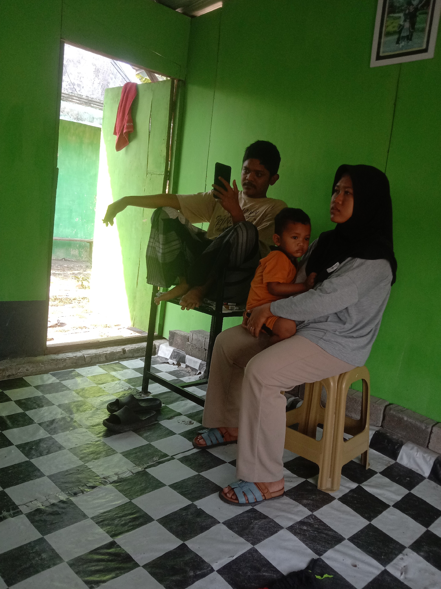 In Kedungrejo, Indonesia, a family is gathered in a room with vibrant green walls. A man, wearing a light-colored t-shirt and plaid pants, sits on a high stool and looks at his phone. Beside him, a woman in a grey long-sleeved shirt and a black hijab holds a young child dressed in an orange t-shirt. The child faces forward with a serious expression. The floor is covered in a black and white checkered pattern. A doorway to the left reveals an outdoor scene with sunlight streaming in, illuminating a red towel hanging on a light green door. A framed picture hangs on the far green wall. The scene depicts a moment of casual family interaction within a simple home environment.
