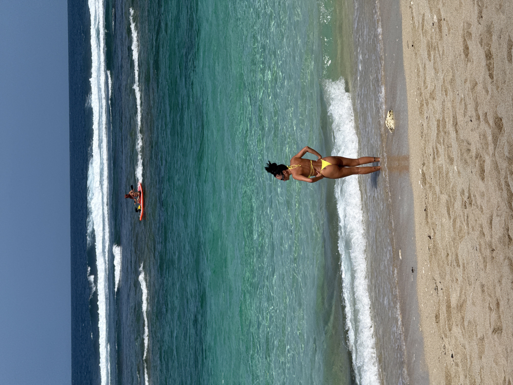 The image shows a woman in a yellow bikini standing at the edge of a beach, looking out towards the ocean. The water is clear and turquoise in color, with waves breaking in the distance. A small orange boat with people in it is visible further out in the water. The beach is sandy and light brown. The sky is blue. The location is likely in Kuta Selatan, Indonesia, given the beach environment and clear water.