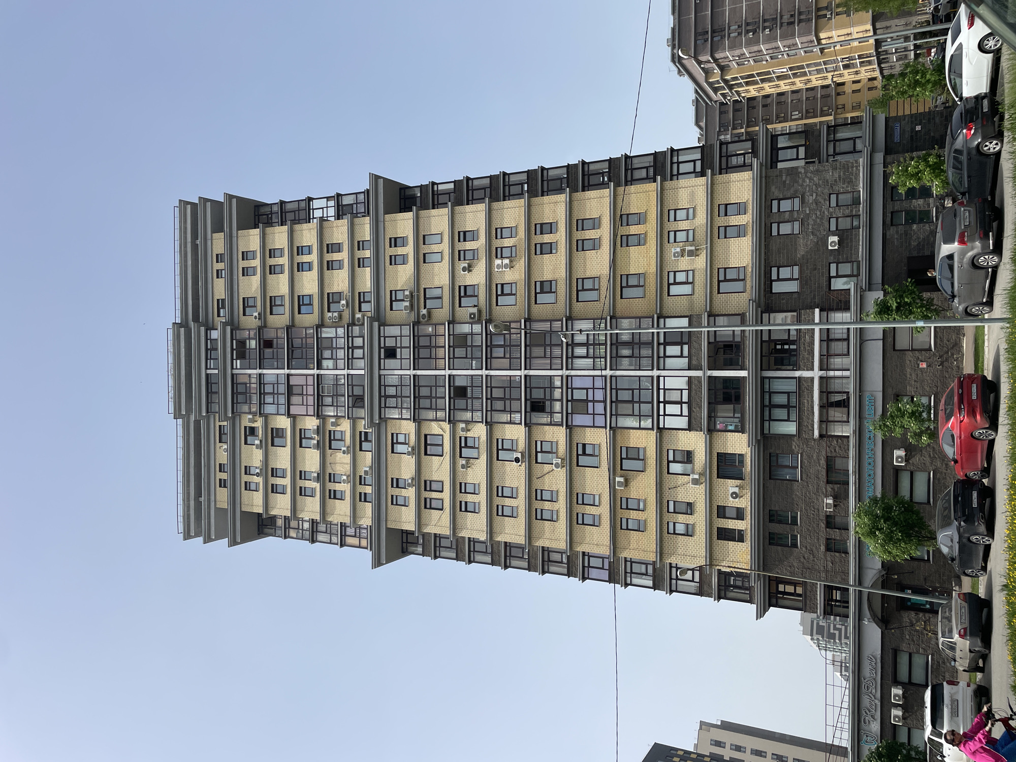 The image shows a tall apartment building in Kazan, Russia. The building has a facade of alternating tan brick and gray stone sections. The windows are dark framed. Air conditioning units are visible in many windows. In the foreground, there are parked cars of varying colors (red, silver, black, white) along the curb. In front of a gray section, there is a sign with Cyrillic text "Ray Dent" and "Стоматологический центр". There are power lines visible against a light blue sky. Adjacent buildings are partially visible on either side of the main building.