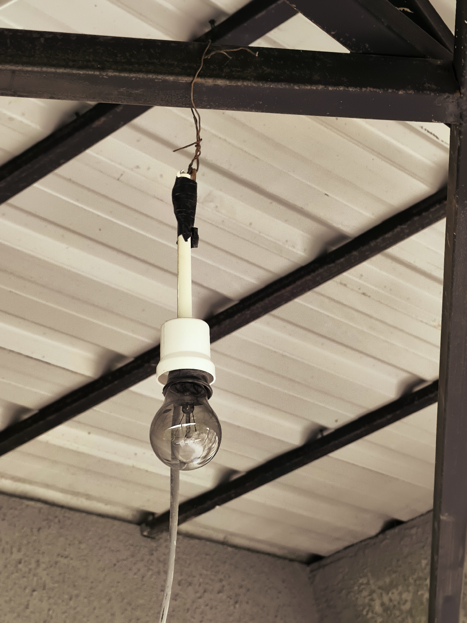 A close-up, low-angle shot captures a bare light bulb hanging precariously from a white plastic socket. The bulb is suspended by a grey wire and an electrical cord. The electrical connections at the top appear to be makeshift, with black electrical tape securing wires to a white pipe, which is then attached to a thin, rusty wire that hangs from a dark metal beam. This beam is part of a larger dark metal structure that supports a corrugated white roof. The roof's ribbed texture is clearly visible, with dark shadows cast between the corrugations. Below the roof and metal structure, a rough, grey concrete wall is visible. The overall impression is one of a hastily installed and somewhat unsafe lighting solution, likely outdoors or in an unfinished space. The specific location within the city of Batumi, Georgia, cannot be determined from the visual information provided in the image.