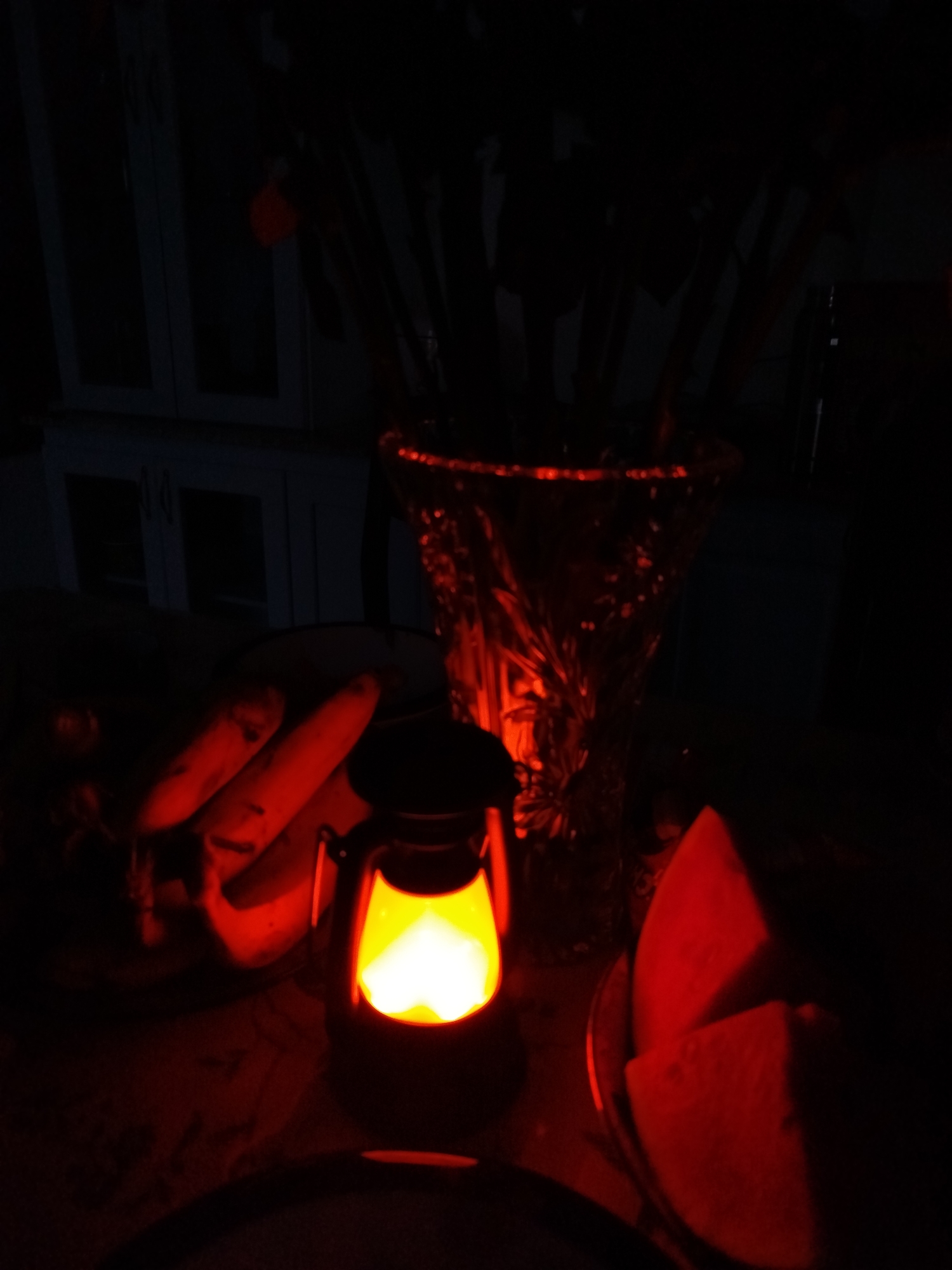 A dimly lit room is illuminated by a central lantern, casting a warm orange glow on the surrounding objects. A decorative glass vase, filled with what appear to be long-stemmed flowers or branches, stands behind the lantern. To the left of the lantern, there are two oblong shapes that resemble fruits or vegetables, possibly bananas. To the right of the lantern, there's what appears to be two slices of watermelon. The objects are arranged on a dark surface, suggesting a table or countertop. In the background, blurred outlines hint at cabinetry or furniture. The overall scene conveys a sense of intimacy and warmth. There is no information to suggest this image was taken in Lạc Dương, Vietnam.