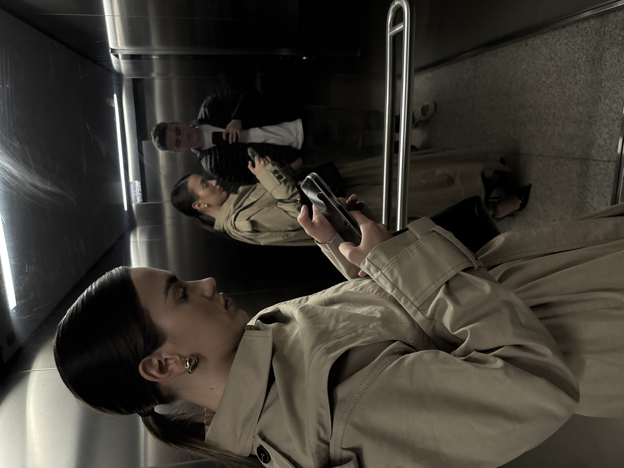 A reflective photograph shows a person wearing a tan trench coat holding a smartphone. The person has dark hair pulled back in a ponytail and is wearing round earrings. In the reflection, another person is seen in a black jacket and white shirt, also holding a phone. The scene appears to be inside an elevator, with metal walls and a vertical handrail visible in the reflection. The location is Unknown, Unknown.