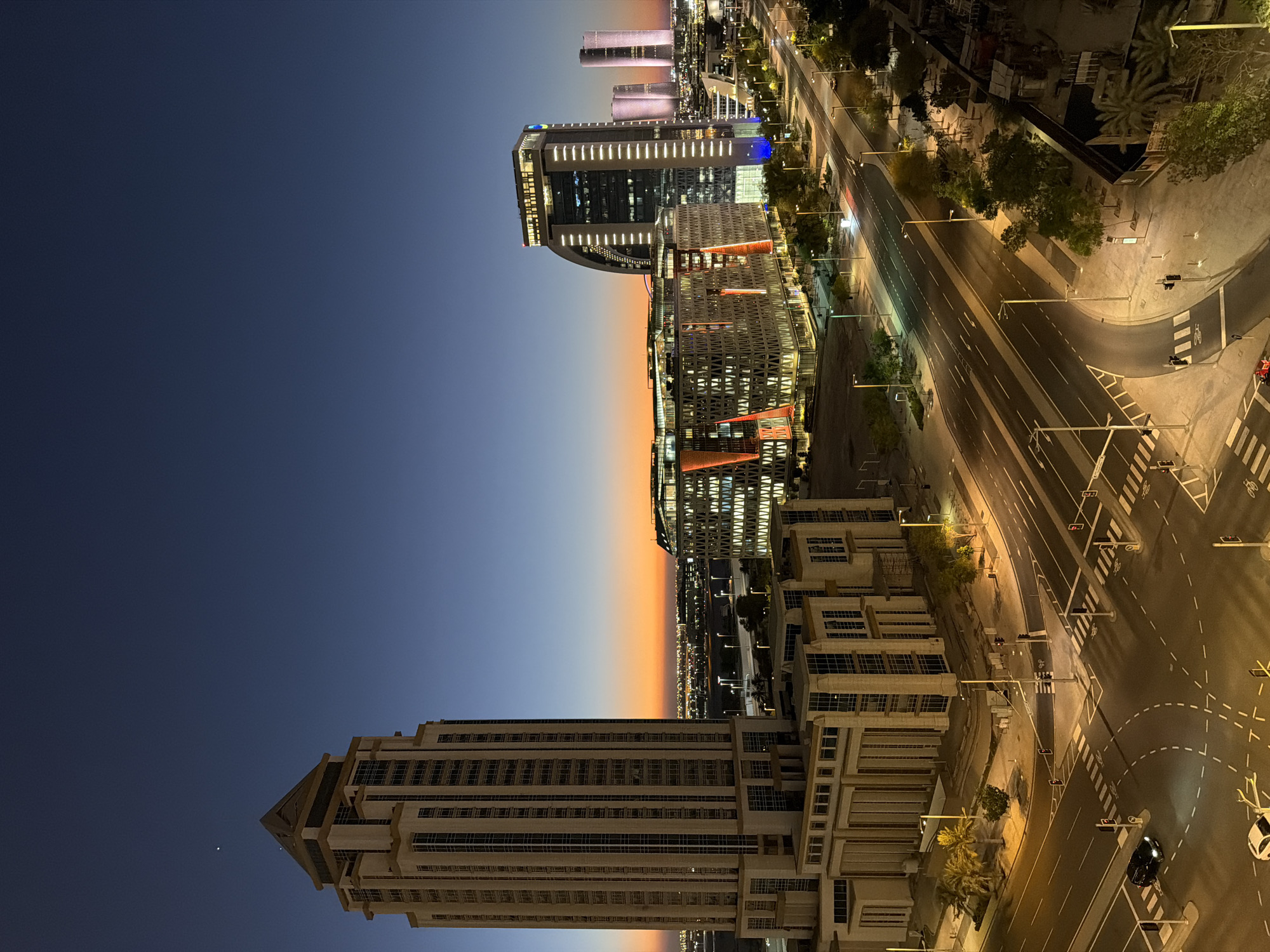 An aerial, horizontal, full shot captures a city skyline at dusk. In the foreground, a tall, light-colored skyscraper with a peaked roof stands to the left. Adjacent to it, on the right, is a modern, wide building with an illuminated facade featuring geometric patterns. Further in the distance, a curved skyscraper with a prominent white stripe illuminates the skyline, flanked by other illuminated skyscrapers. A multi-lane road curves through the scene, with streetlights casting a warm glow. Two cars are visible on the road. The sky transitions from a deep blue at the top to a fiery orange near the horizon, indicating either sunrise or sunset. The scene suggests a developed urban environment with contemporary architecture.