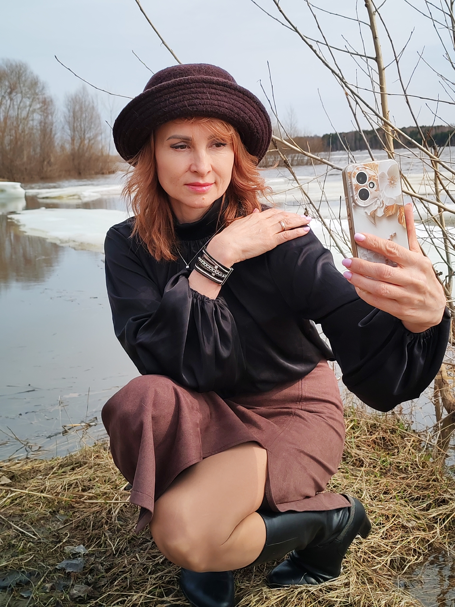 A middle-aged woman is crouching near a body of water partially covered in ice, possibly in the vicinity of Поварёнки, Russia. She is wearing a brown hat, a long-sleeved black blouse with puffy sleeves, a brown skirt, and black boots. She is holding a smartphone in her right hand, apparently taking a selfie. She is wearing a bracelet on her left wrist and a ring on her left hand. The background includes a body of water with patches of ice, bare trees, and some vegetation. The ground where she is squatting is covered in dry grass. The sky is overcast.