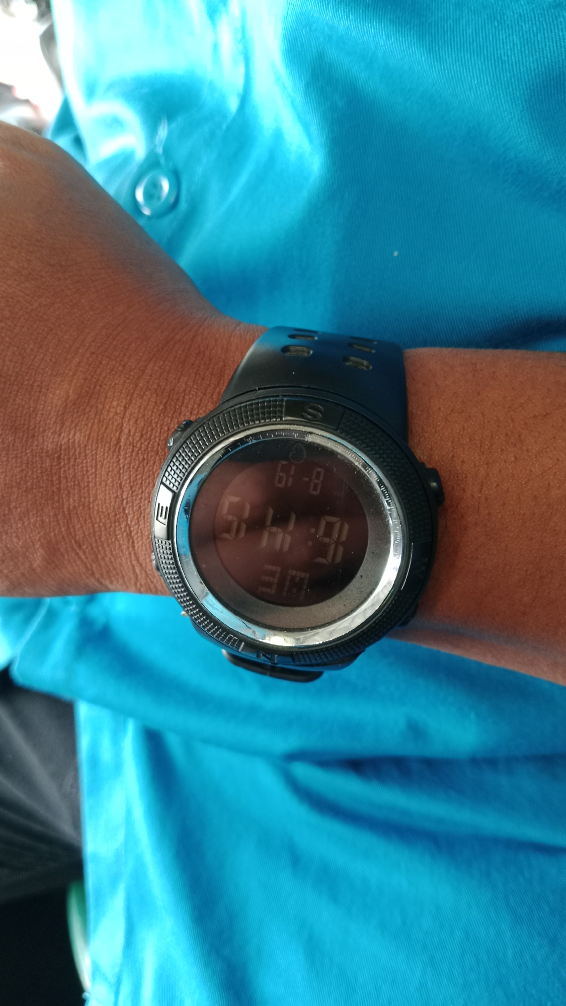 A close-up shot shows a black digital watch on a person's wrist. The watch displays the time as 16:14:19 and the date as 8-19. The watch is worn on a person's arm, which is covered by a blue shirt. The background is out of focus but appears to be a light blue fabric. The watch has a round face with a black bezel and silver accents. The band of the watch is also black and appears to be made of rubber or silicone. The image is well-lit and the details of the watch are clear. This image does not provide enough information to determine its location in the city of Handil Bakti, Indonesia, or any other specific location.