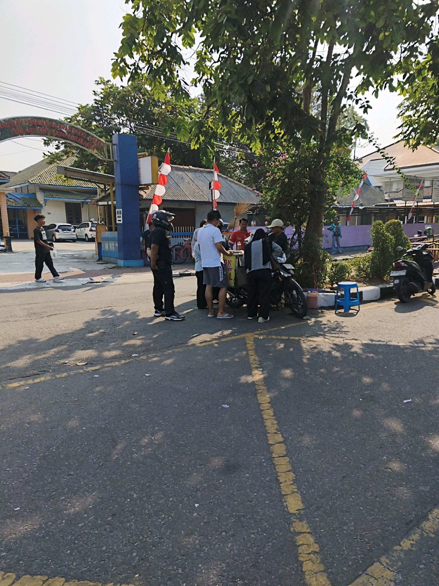 The image shows a street scene in Surakarta, Indonesia, on a sunny day. Several people are gathered around a food cart, and two motorcycles are parked nearby. In the background, there are buildings, trees, and Indonesian flags. The overall atmosphere appears to be casual and relaxed.
The scene is set on a street with asphalt pavement and yellow painted lines. The food cart is positioned under a tree, providing shade. The people around the cart seem to be customers and possibly vendors. One person is wearing a helmet, suggesting they may have arrived by motorcycle. The buildings in the background are typical of Indonesian architecture, with tiled roofs. The presence of Indonesian flags indicates a patriotic or celebratory occasion. The sunlight casts shadows on the ground, indicating the time of day.