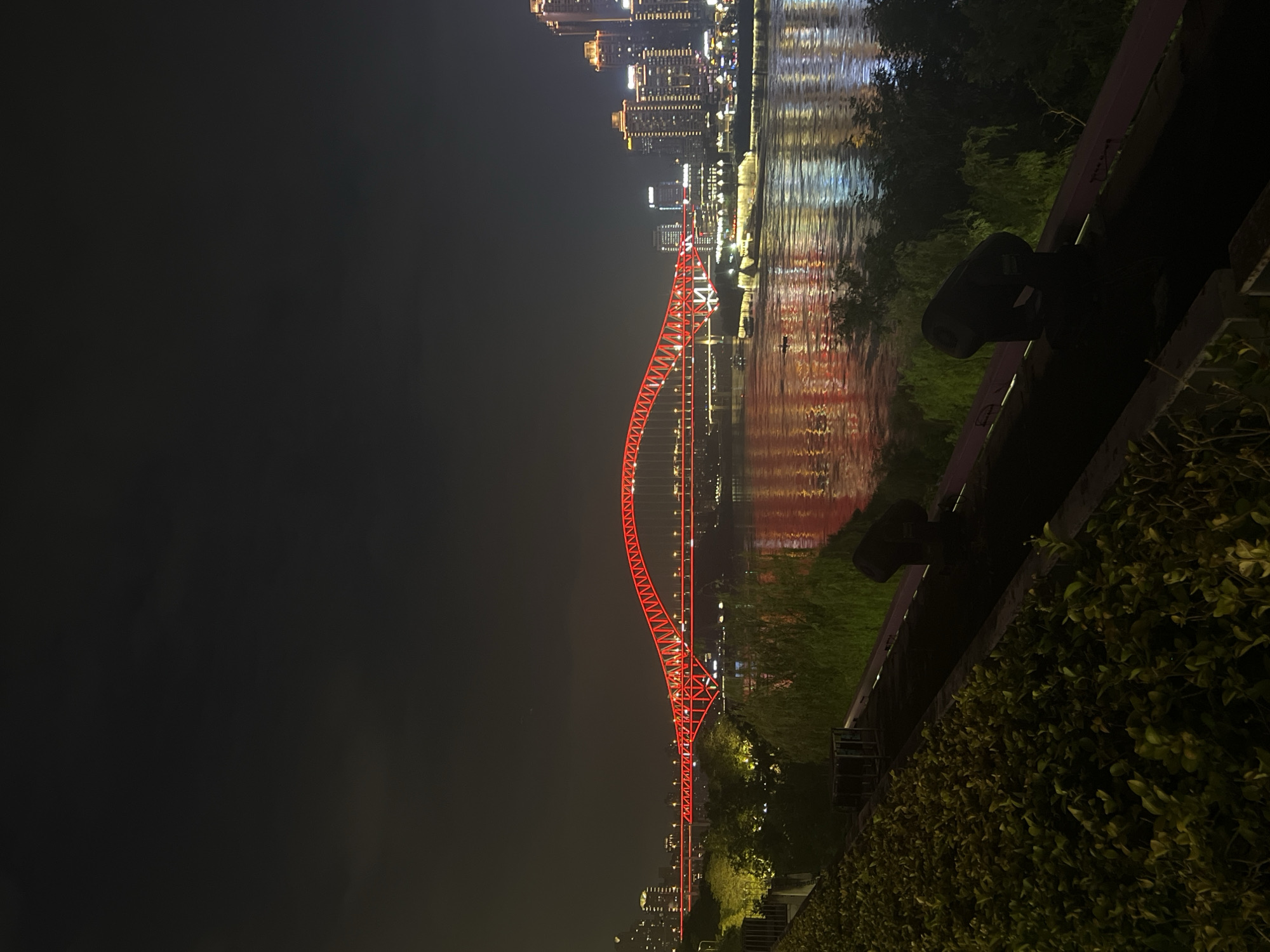 This is a nighttime photograph of the Jialing River in Jiangbeicheng Street, China, taken from a vantage point overlooking the water. A prominent red illuminated bridge, possibly the Jialingjiang Bridge, spans the river, its vibrant lights reflecting on the water's surface. On the far bank, a cityscape of illuminated skyscrapers and buildings forms a dense urban backdrop. The foreground features a landscaped area with lush green foliage and several dark, cylindrical spotlights positioned on a raised platform. The overall scene captures the illuminated, modern architecture of the city at night, contrasted with the dark water and foreground vegetation.