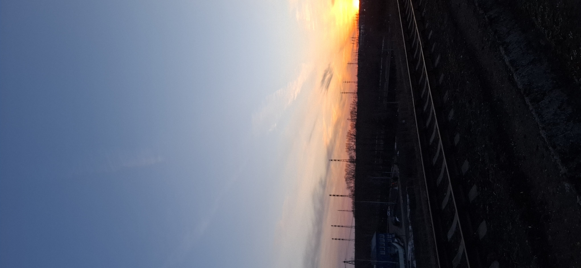 The image shows a sunset over a railway line in Chaikovsky, Russia. The sky transitions from blue on the left to orange and yellow near the top of the frame, indicating the setting sun. Silhouetted against the sunset are power lines and trees. To the right of these, the railway tracks run diagonally across the frame. The area around the tracks is dark. Buildings are visible in the lower right of the image.