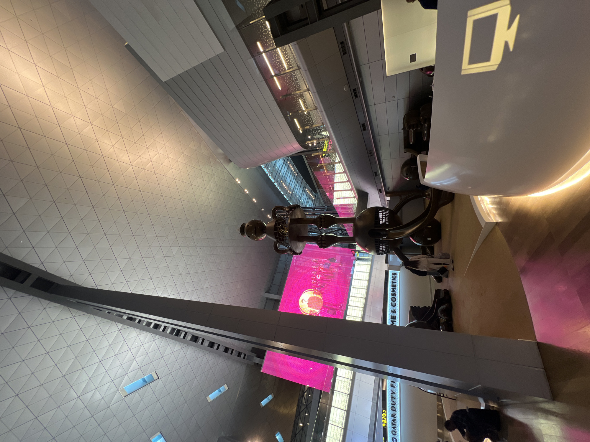 This image captures a large, modern indoor space, likely an airport terminal, characterized by its expansive architecture and contemporary art installation. The most prominent subject is a large, polished metallic sculpture of a stylized bear-like figure, standing centrally with its head tilted slightly. This figure has a somewhat cartoonish appearance with a round head, a stout body, and slender limbs.
The setting features a high ceiling with intricate geometric patterns made of triangular panels, suggesting a sense of depth and modern design. Below, the space is multi-level, with walkways or mezzanines supported by sturdy beams, and glass railings providing views across the different levels. Large, vibrant pink digital screens are visible in the background, one displaying what appears to be a golden coin or medal with a face on it.
Several individuals are scattered throughout the scene, either walking, standing, or in one instance, squatting near the bottom right. Their presence suggests normal activity within a public area. To the right, there's a white counter or kiosk with a distinct symbol resembling a stylized video camera or screen projected or illuminated on its surface.
Visible text provides strong location cues: "QATAR DUTY FREE FI" (likely "QATAR DUTY FREE FASHION") and "ME & COSMETICS" (likely "PERFUME & COSMETICS"), indicating a commercial area, most probably within Hamad International Airport in Doha, Qatar, where the "Lamp Bear" sculpture is a famous landmark. The scene is brightly lit by artificial light, making the time of day indistinguishable.