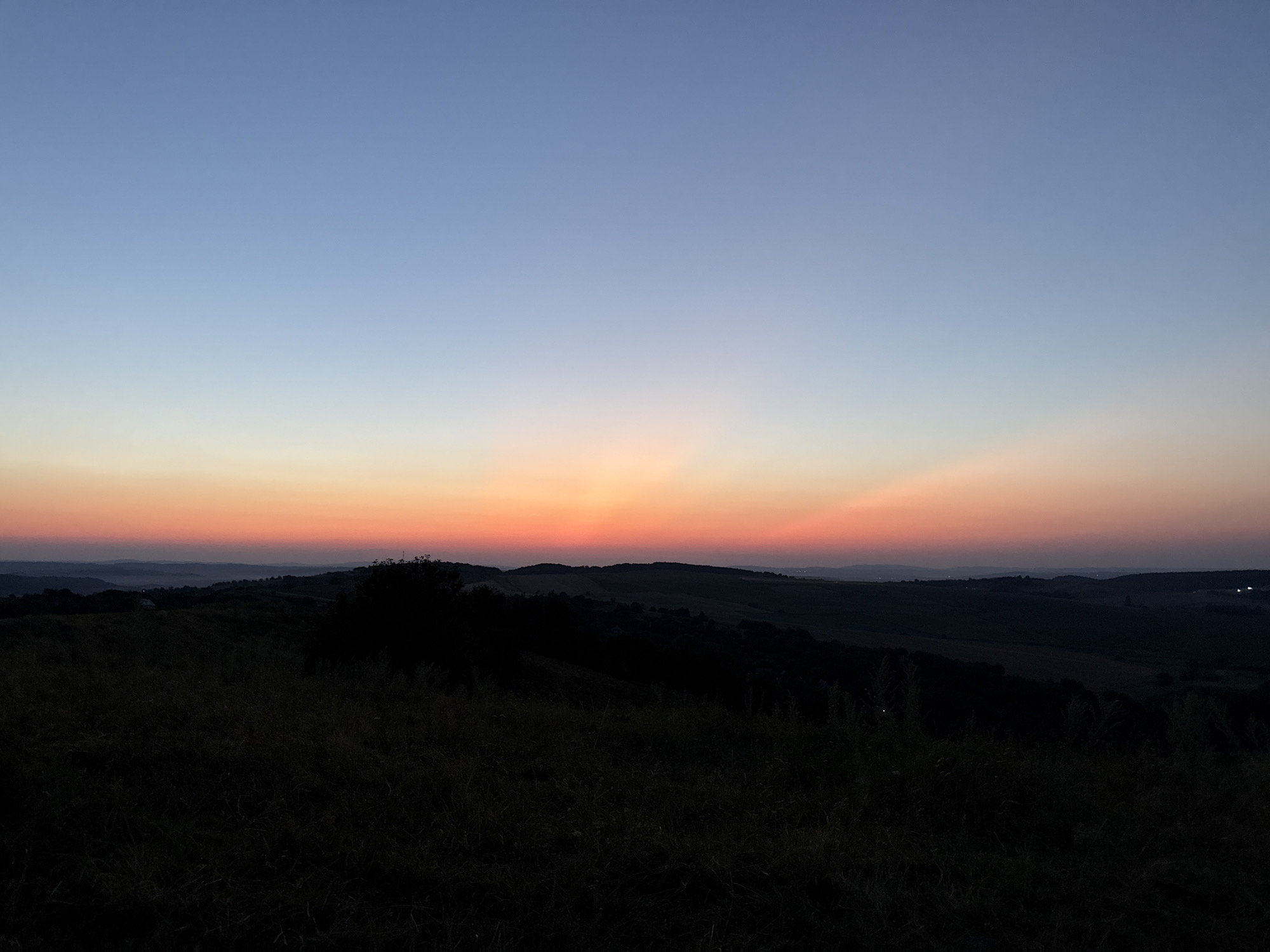This is a photograph of a sunset over a hilly landscape. The sky is a gradient of colors, from a light blue at the top to orange and pink near the horizon. The sun's rays are visible, fanning out from the horizon. In the foreground, there is a silhouetted grassy field. In the middle ground, there are rolling hills covered in trees and vegetation, also in silhouette. In the distance, there are faint lights visible, suggesting a town or city. The overall mood of the image is peaceful and serene.