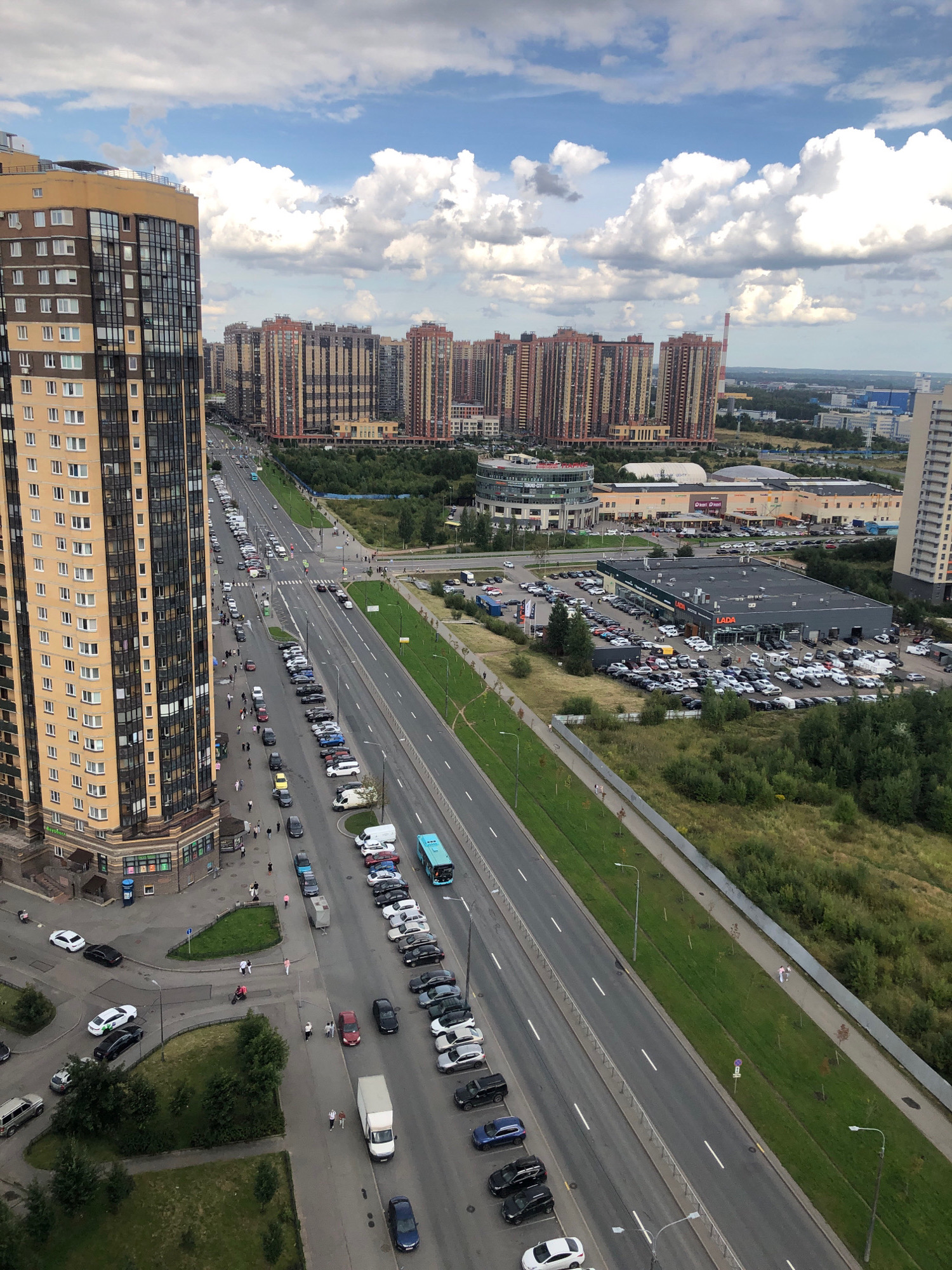 The aerial view captures a modern urban landscape in St. Petersburg, Russia, on a cloudy day. A prominent, tall residential building with a yellow facade and dark windows stands on the left, dominating the foreground. Adjacent to it, a multi-lane road runs vertically through the image, lined with parked cars and a few pedestrians. Across the road, a commercial area features large, multi-story buildings with similar architectural styles, including what appears to be a car dealership with "LADA" branding. Beyond these structures, more residential complexes rise in the distance, characterized by their colorful facades. Green spaces, including trees and grassy areas, are interspersed throughout the scene, with a distinct patch of undeveloped land on the right. The sky is filled with dramatic white clouds against a blue backdrop, indicating partly cloudy weather. The scene suggests a bustling urban environment with residential, commercial, and transportation infrastructure.