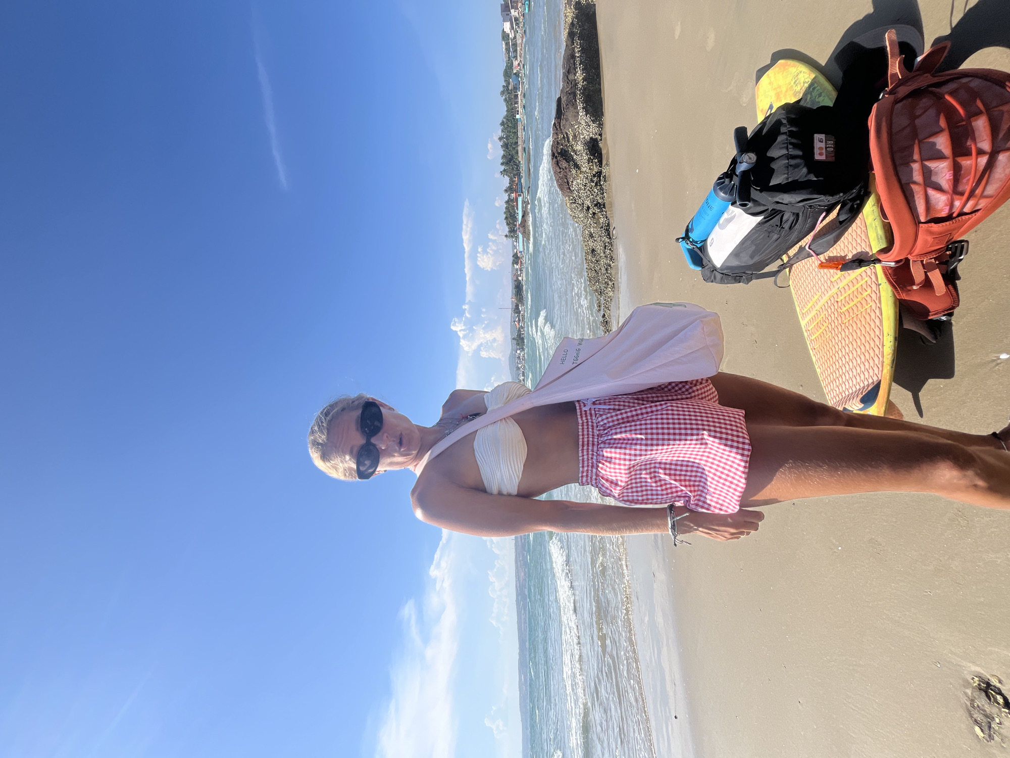 A woman with blonde hair pulled back and wearing dark sunglasses stands on a sandy beach in Mũi Né, Vietnam. She is wearing a white strapless bikini top and red and white checkered shorts. A pink canvas bag with the text "HELLO MESSAGE" is slung over her left shoulder. In the background, the ocean has moderate waves, and a line of buildings and palm trees stretches along the coastline. To the woman's right, two kiteboards are propped up, one orange and yellow and the other yellow. A black backpack with a blue water pump is strapped to the top kiteboard, and a red kiteboarding harness is placed on the front of the lower kiteboard. The sky is a clear, bright blue with a few scattered white clouds. The sand is wet near the water's edge and drier further up the beach.