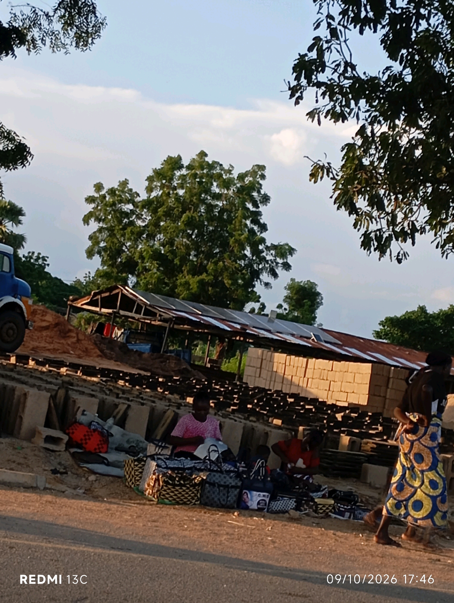 In Shendam, Nigeria, this image captures an outdoor market scene with a focus on a stall displaying an array of bags. Several individuals are present, including at least two women and a child actively engaged in selling or arranging merchandise. The market is situated near a construction area, evident from stacks of building materials like bricks and a partially constructed shelter with a corrugated metal roof. A blue truck is parked on the left side of the frame, suggesting logistics or transport related to the market or construction. The overall environment appears to be a roadside market, with trees and a partially cloudy sky indicating it is daytime. The lighting suggests it might be late afternoon or early evening.