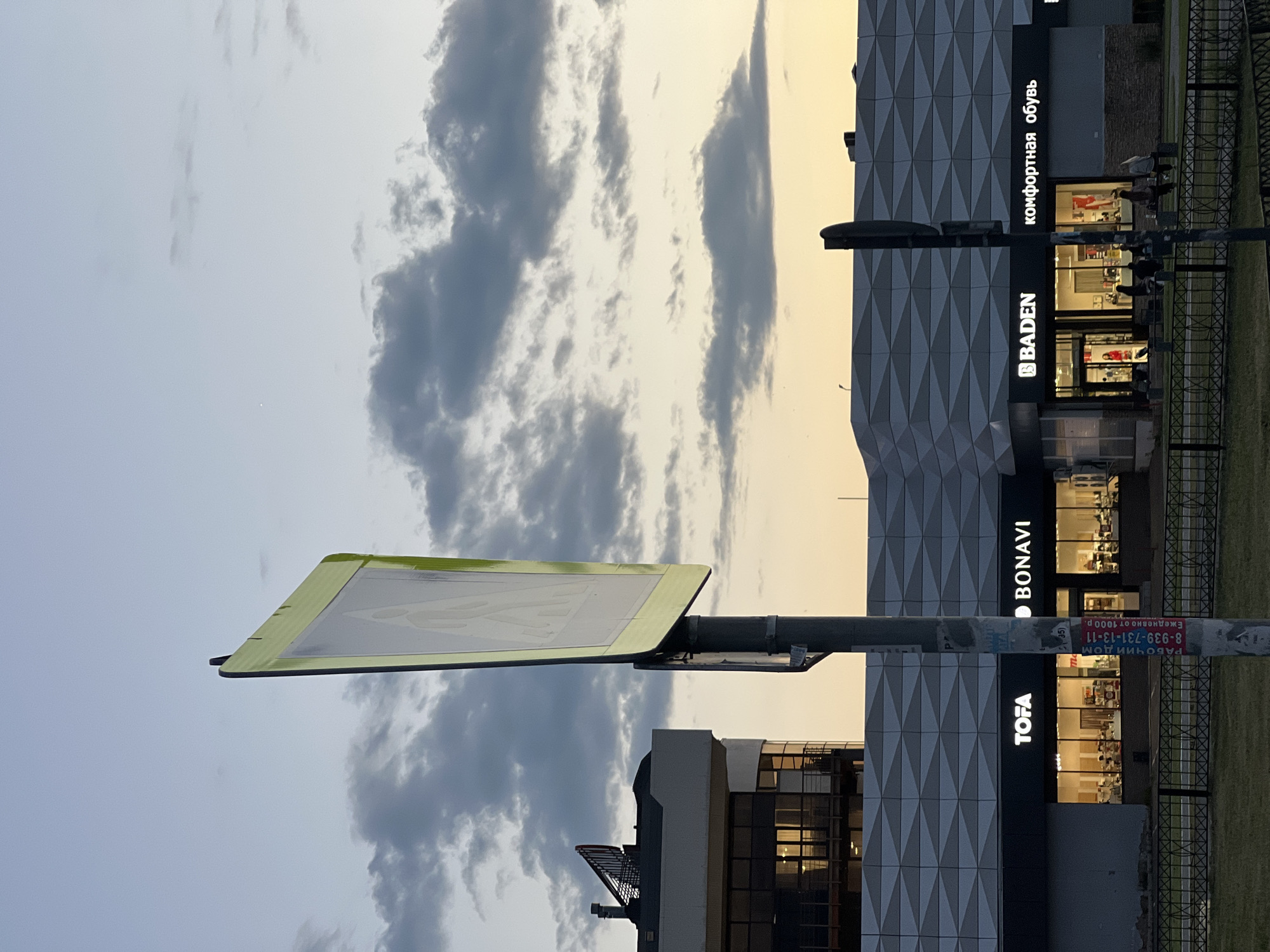The image depicts a pedestrian crossing signpost in the foreground, with a building complex featuring multiple storefronts in the background. The sky above is overcast with grey clouds, transitioning to a lighter hue near the horizon. The building's facade is modern, with a textured pattern and illuminated signage.
Specifically, the signs visible on the building indicate the presence of stores such as "TOFA", "BONAVI", and "BADEN". The "BADEN" store also has signage that reads "комфортная обувь," which translates to "comfortable shoes." The building is located in Kazan, Russia, as indicated by a sticker on the signpost that reads "РАБОЧИЙ ДОМ 8-939-731-13-11 Ежедневно от 1000 р." which translates to "WORK HOUSE 8-939-731-13-11 Daily from 1000 p."
In front of the building, a black metal fence runs along a grassy area. Several people are visible seated on benches and walking near the storefronts, suggesting a public space. A multi-story building with a contemporary design, featuring large windows and a red-capped structure on its roof, is situated to the left of the main building complex. The overall scene appears to be captured during the evening or late afternoon, given the ambient light.