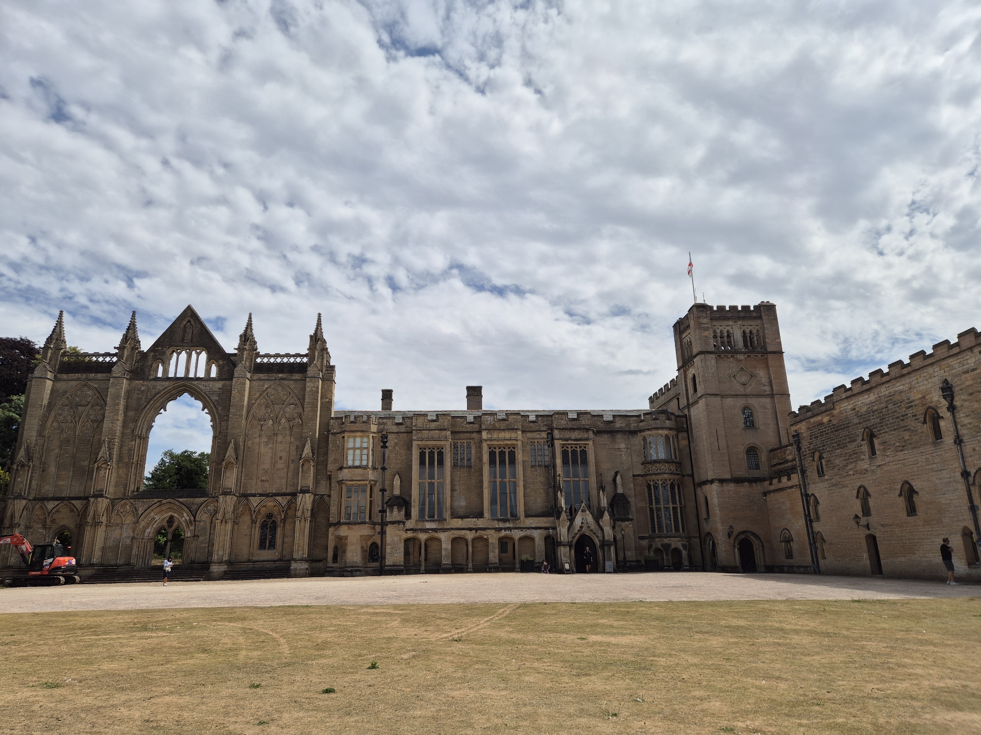 The image shows the exterior of a large, historic building with stone architecture, likely a castle or abbey, under a cloudy sky. The building features Gothic-style arches, windows, and a prominent tower with a flag. In the foreground, a dry, grassy courtyard is visible, with a few people present. An excavator is parked in front of one of the ruined sections of the building, suggesting ongoing restoration or maintenance work. The building appears to be a tourist attraction or a historical site. This is Wollaton Hall in Nottingham, United Kingdom.