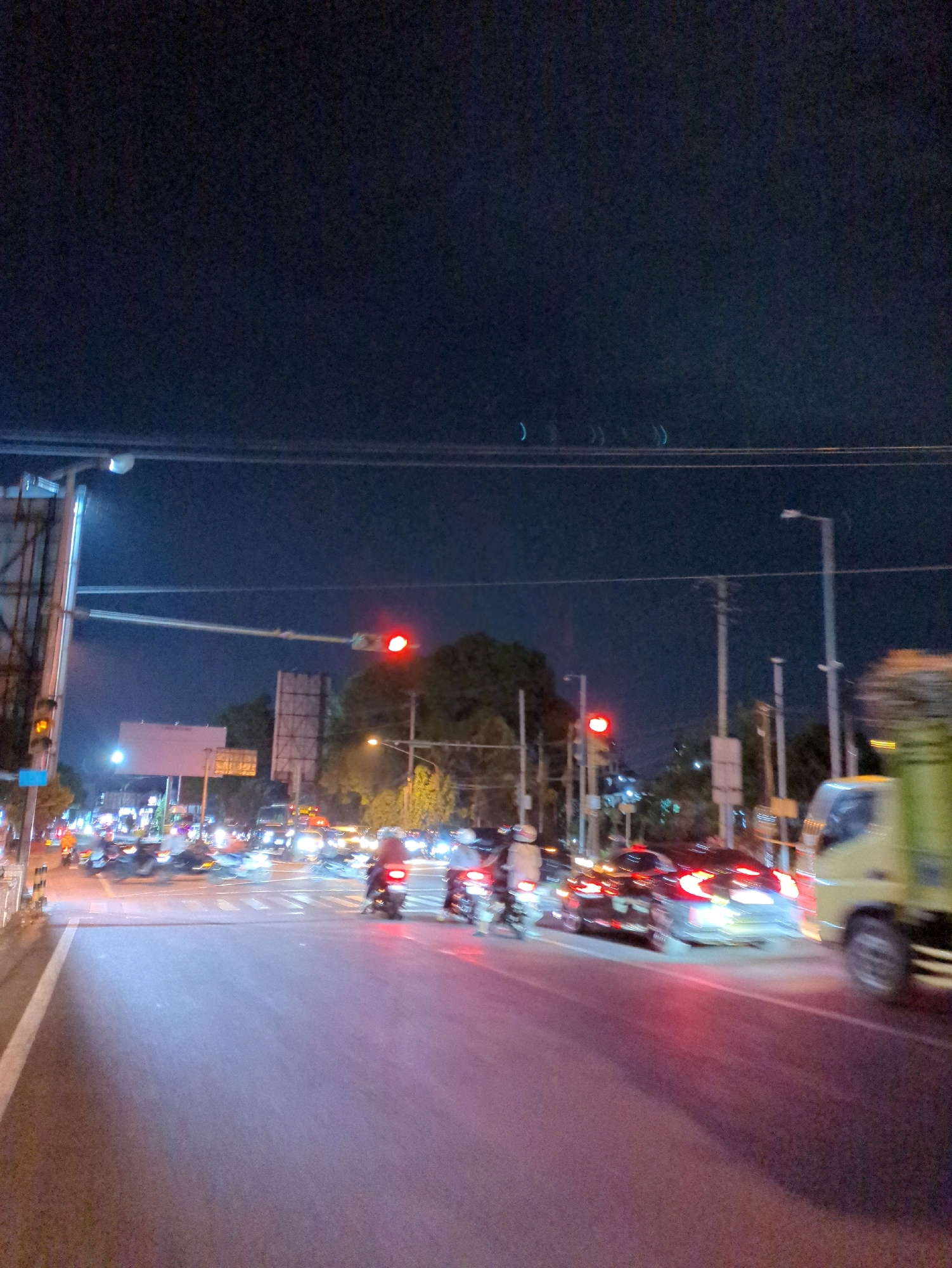 The image depicts a nighttime scene of a busy intersection in Surakarta, Indonesia. Traffic is moving, with cars and motorcycles visible, some with their lights blurred due to motion. A yellow truck is present on the right side of the frame. Traffic lights are illuminated, with a red light prominently displayed on the left. The sky is dark, indicating it is night. The road surface is asphalt, and there are streetlights and overhead wires visible. Buildings and trees are present in the background, illuminated by the surrounding lights. The overall impression is a dynamic urban environment at night.