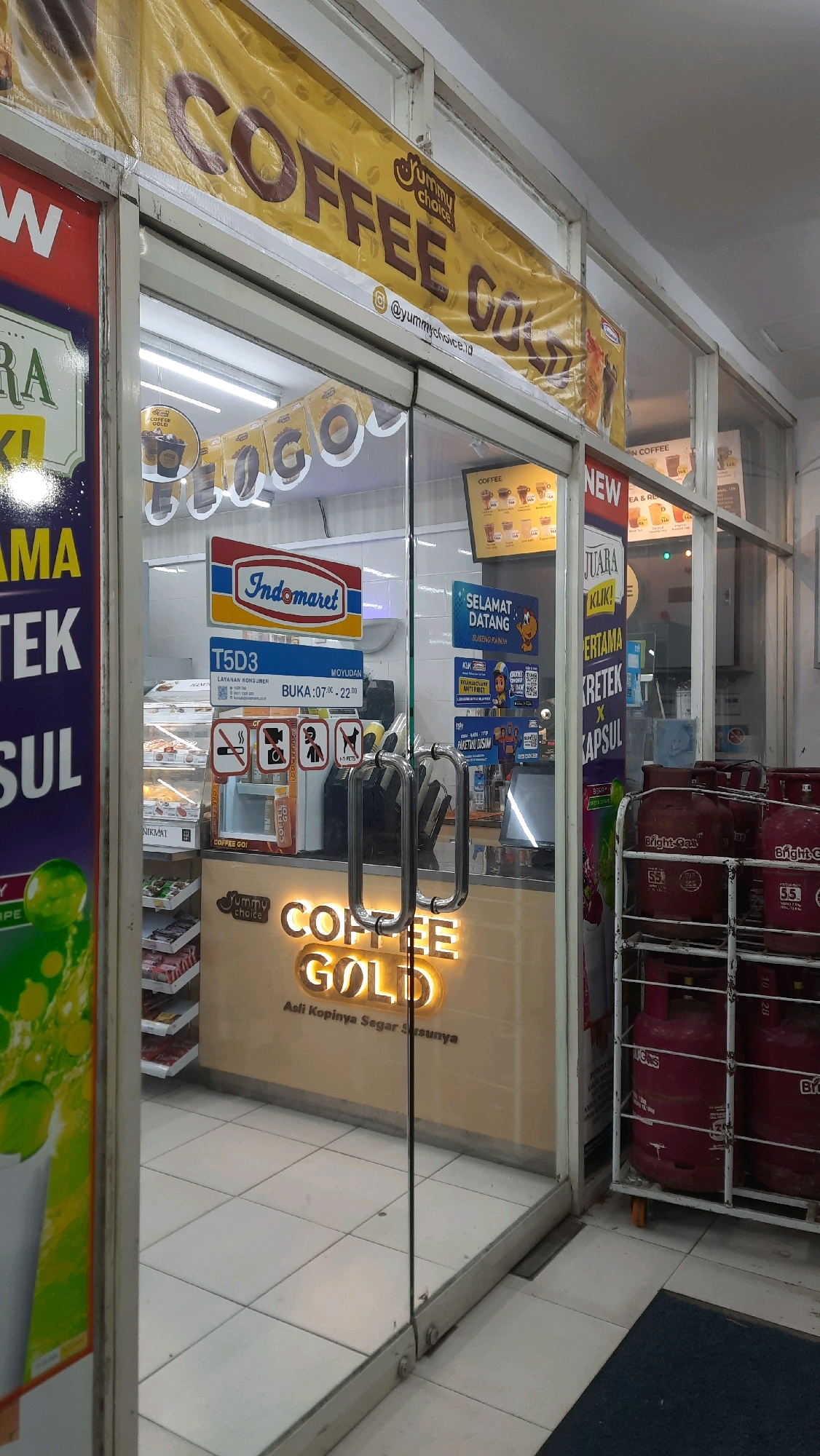 This is a medium shot of the exterior of a Yummy Choice Coffee Gold store, likely a franchise located within an Indomaret convenience store in Moyudan, Indonesia. The storefront features large glass doors, through which the interior can be seen. A yellow banner with "COFFEE GOLD" in large brown letters is prominently displayed above the entrance, with the Yummy Choice logo and social media handle "@yummychoice.id" visible.
Inside the store, there are shelves stocked with products, and a counter area where coffee is likely prepared and sold. Various signs are visible, including an "Indomaret" logo, a "SELAMAT DATANG" (Welcome) sign, and operational hours for "T5D3 LAYANAN KONSUMEN" which are 07:00 to 22:00. Images of coffee drinks are displayed on a menu board. To the right of the entrance, several red LPG gas cylinders are stacked on a metal rack. The floor is tiled with a light-colored grid pattern. The overall scene depicts a retail environment.