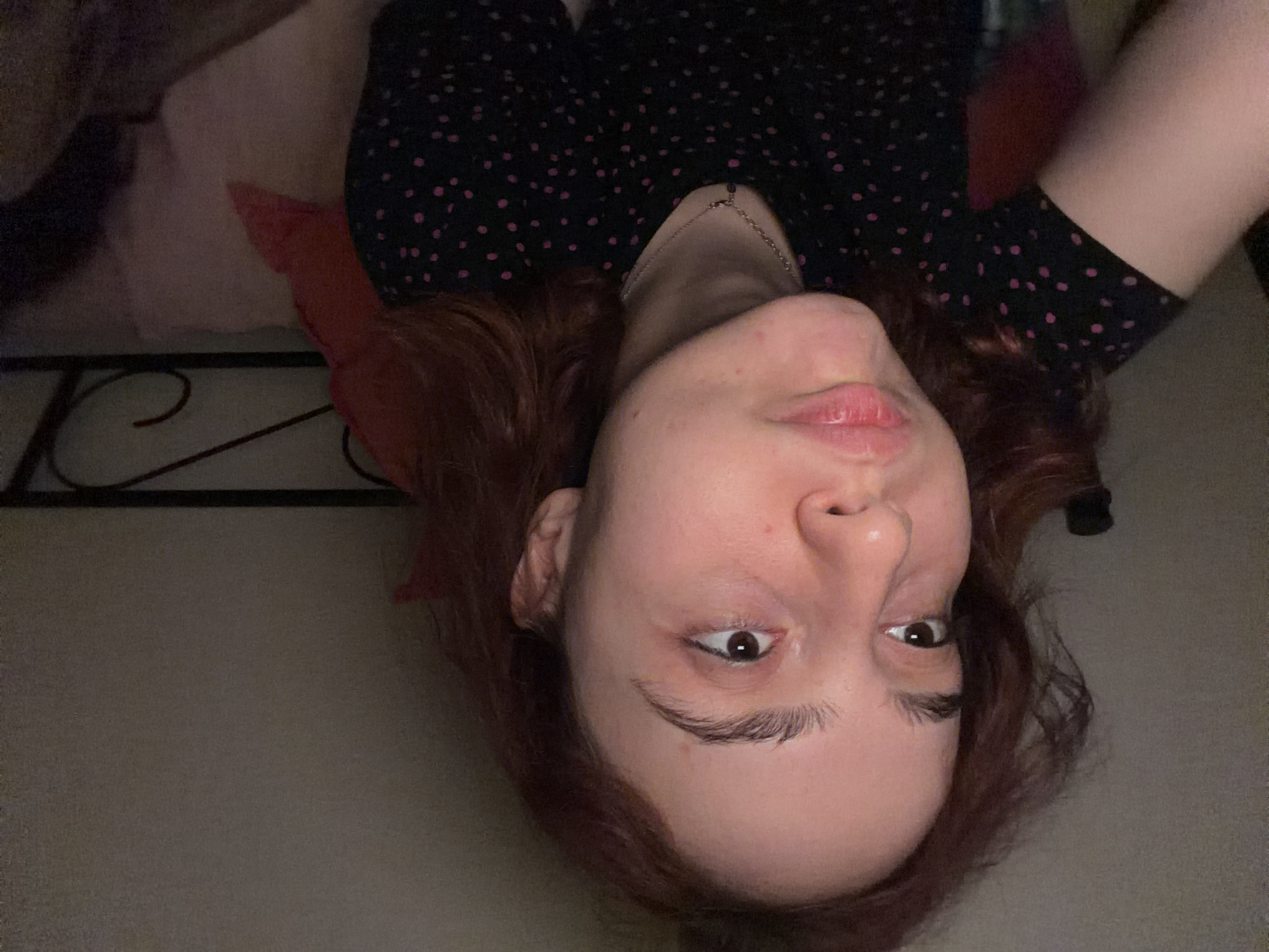 The image shows a close-up portrait of a young woman with reddish-brown hair, looking to her left with her cheeks puffed out and lips pursed. She is wearing a black t-shirt with a pink polka dot pattern and a delicate silver necklace. The background is dimly lit and appears to be indoors, with a dark wall behind her. A section of a metal bed frame and pillows are visible to her right. There is no indication of Strasbourg, France in the image.