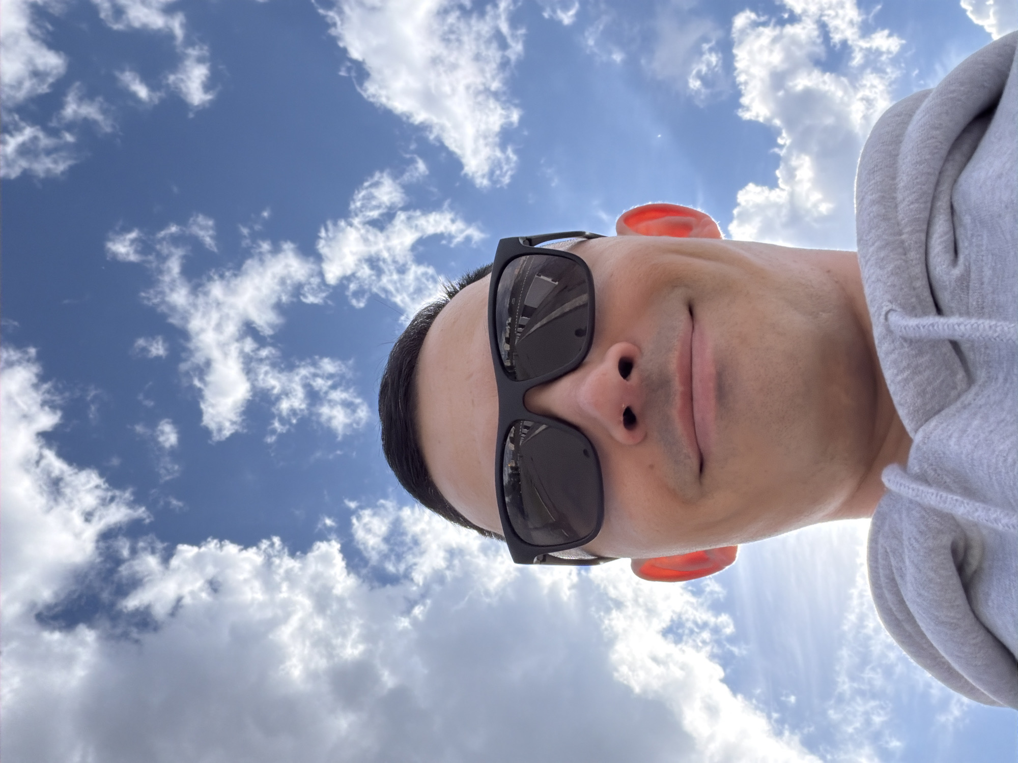 A low-angle, distorted selfie shows a person wearing black-framed sunglasses and a gray hooded sweatshirt against a backdrop of a blue sky with scattered clouds. The person's face is positioned in the lower portion of the frame, with the sky and clouds taking up most of the background. The reflection in the sunglasses suggests an urban environment with buildings, possibly taken in Izhevsk, Russia. The person's ears are visible.
