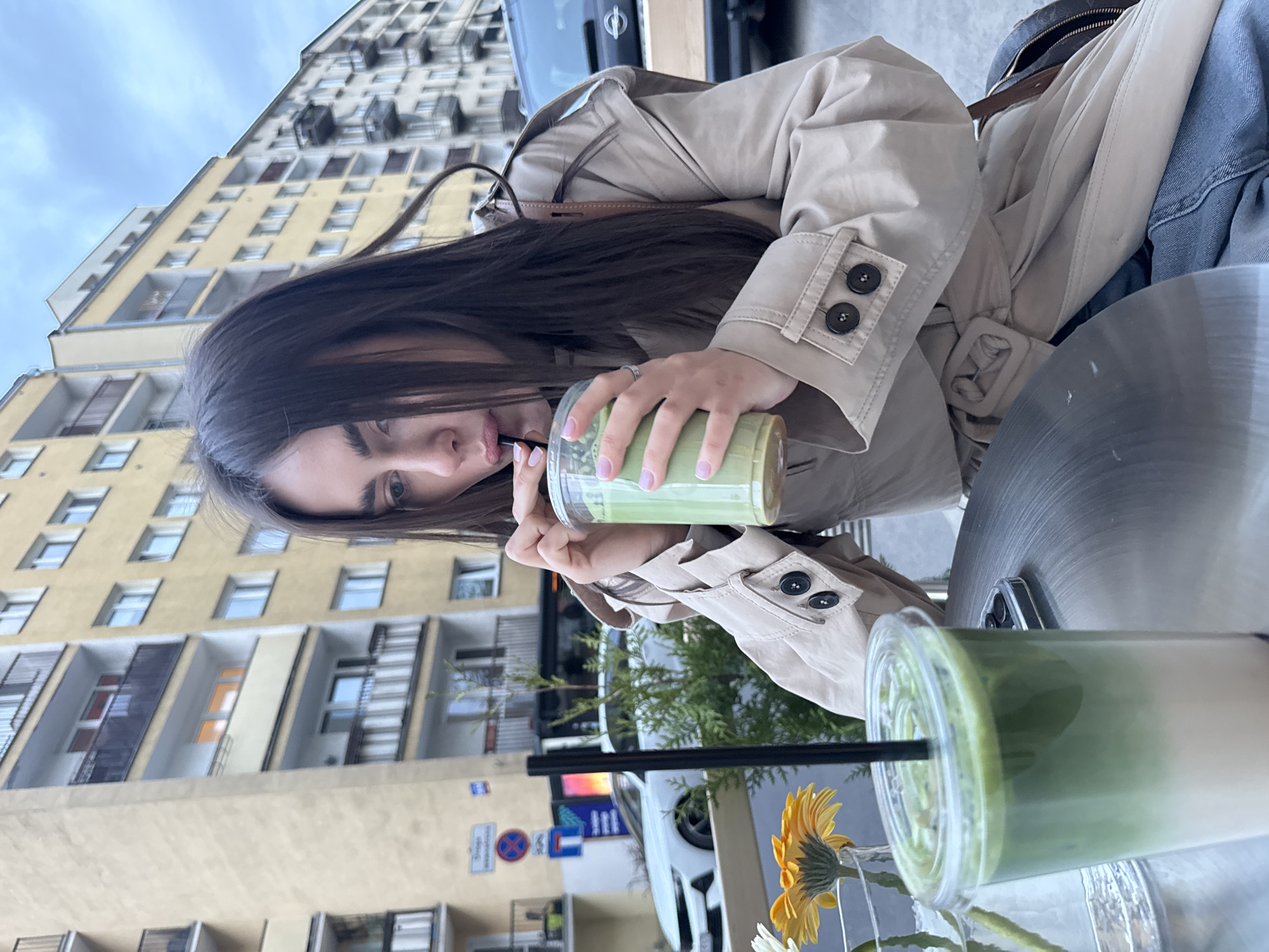 The image, rotated 90 degrees clockwise, captures a woman enjoying a drink outdoors in an urban setting.
**Subjects:** The central subject is a young woman, seen in a diagonal composition from the chest up. She has long, dark hair that partially frames her face as she looks towards the left of the frame. She wears a light beige or khaki trench coat. Her left hand, adorned with light-colored nail polish and a ring, holds a clear plastic cup containing a layered green and white beverage, likely a matcha latte, from which she sips through a black straw. Another identical drink in a clear cup with a black straw is visible on a metallic-looking table in the bottom right corner. Beside it, a small vase holds a yellow daisy-like flower and white flowers. In the upper right quadrant, parts of a brown bag strap and blue denim fabric (possibly a jacket or jeans) are visible.
**Scene and Setting:** The background features a tall, light yellow or beige multi-story building with numerous windows and balconies, dominating the upper left side of the frame. The sky, partly cloudy and light blue, fills the space behind the building. Below the building, elements of a city street are visible, including parked cars (a dark-colored one partially visible in the upper background, and a white one further back), and various street signs. Green foliage from plants or trees can be seen near the bottom center.
**Activity:** The woman is engaged in the simple activity of drinking a beverage, suggesting a moment of relaxation or a break at an outdoor cafe or street-side establishment.
**Notable Details:** The image's orientation is rotated clockwise. The lighting suggests daytime, possibly under an overcast sky, as shadows are soft and diffuse. One window in the background building shows a warm light. The architecture of the building and the street signs (a circular red-bordered sign indicating "no stopping/standing" and a blue rectangular sign with white, likely Polish, text) provide strong contextual cues for Warsaw, Poland, consistent with the provided location information. The woman's visible eye conveys a neutral or calm expression.
**Visible Text:** A red-bordered circular street sign with a diagonal line (an international "no stopping" or "no standing" symbol) is clearly visible. A blue rectangular street sign with white, partially legible text is also present, consistent with local Polish signage.