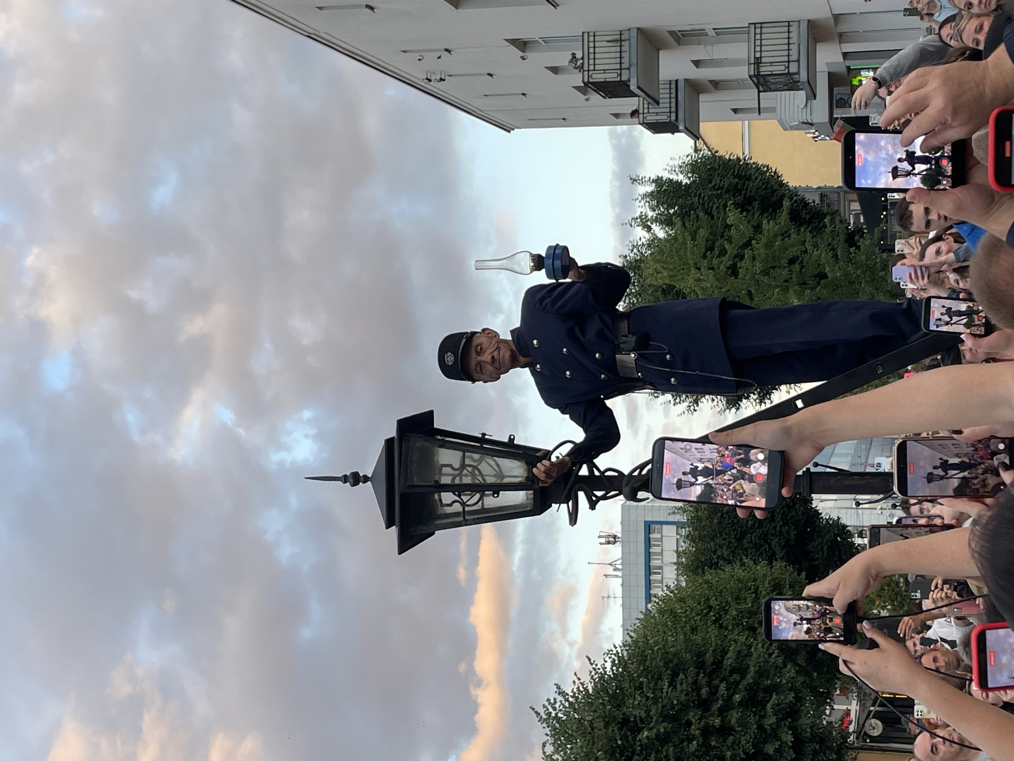In an outdoor urban setting, likely a street in Brest, Belarus, a man dressed as a historical lamplighter stands atop a lamppost. He is wearing a dark blue uniform with gold buttons and epaulets, a cap, and a belt with pouches. He holds a glass kerosene lamp in his right hand and is reaching up with his left hand towards the lantern of the lamppost. Below him, a crowd of people are gathered, many of them holding up smartphones to take pictures or record videos of the man and the lamppost. The lamppost itself is ornate and black, with a large glass lantern at the top. In the background, there are trees, buildings with balconies and windows, and a cloudy sky with hints of pink from the setting sun. One of the buildings in the background features a flag that appears to be the flag of Belarus. The scene suggests a performance or historical reenactment taking place in a public space.