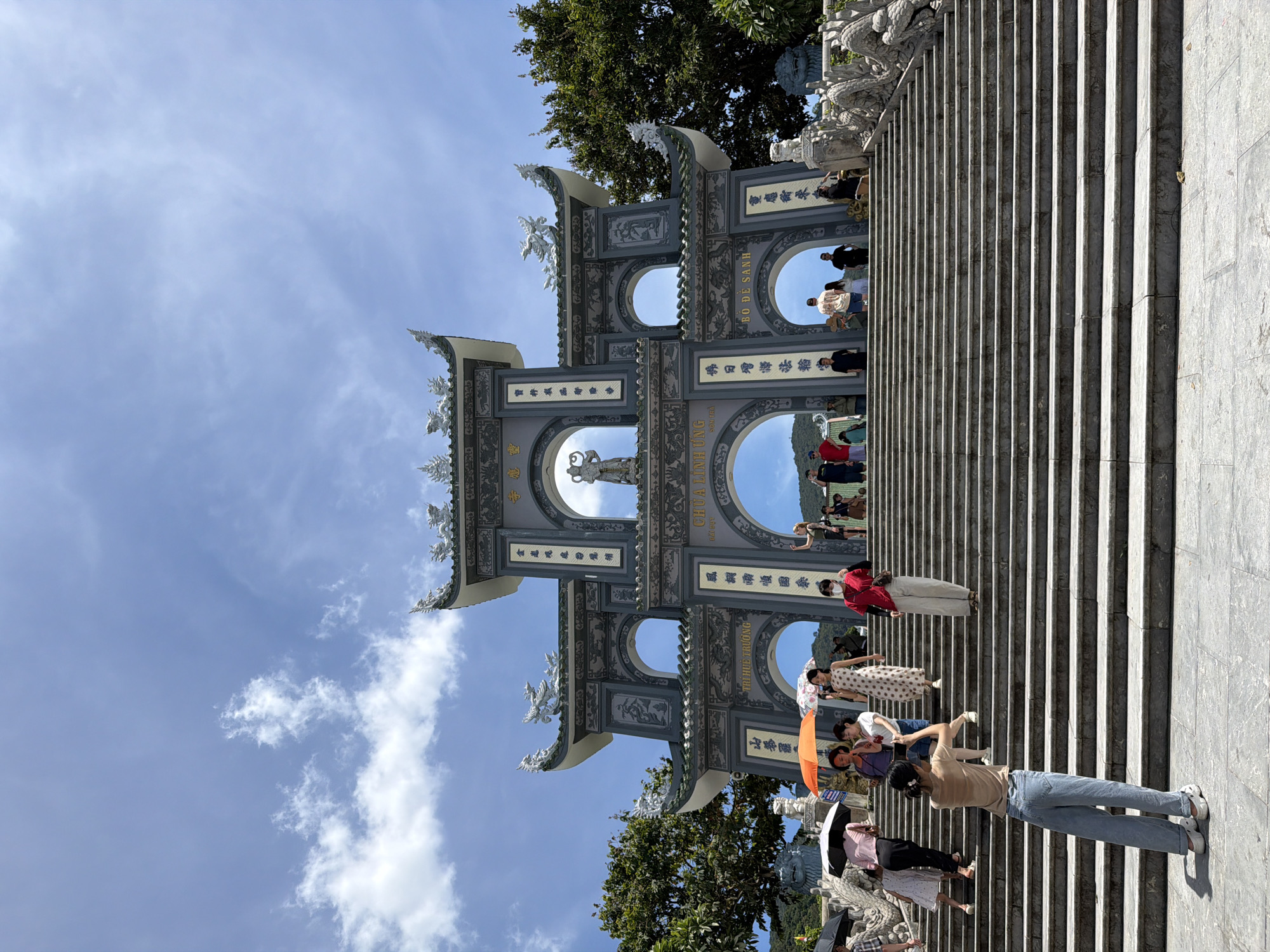 In Đà Nẵng, Vietnam, a wide stone staircase ascends towards a grand, ornate gate with multiple arches, characteristic of traditional Vietnamese temple architecture. The gate, adorned with intricate carvings and dragon motifs, is painted in shades of gray and green. On the upper archway, a statue of a deity is visible. Numerous people are ascending and descending the stairs, some holding umbrellas against the bright, partly cloudy sky. A woman in the foreground, facing away from the camera, is taking a photograph with her phone as she stands on the lower steps. The scene conveys a sense of cultural tourism and spiritual pilgrimage.