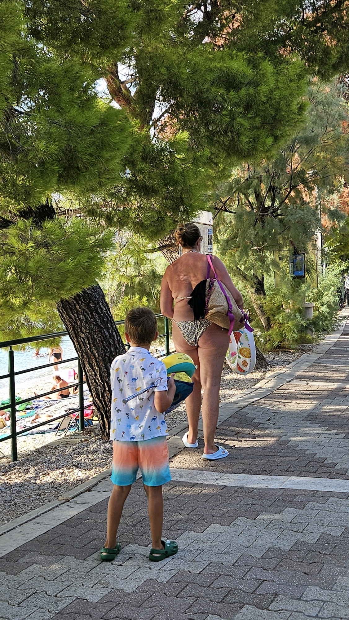 In Općina Tučepi, Croatia, a young boy walks away from the camera on a paved walkway beside a beach. He is wearing a white t-shirt with a dinosaur print, orange and blue ombre shorts, and greenCrocs. He carries a blue and yellow inflatable ball. To his right, a woman walks away from the camera wearing a leopard print bikini and carrying a large patterned tote bag over her shoulder. She has a black backpack slung over her shoulder. The background shows a beach with people sunbathing and the Adriatic Sea. Trees with green foliage line the walkway.