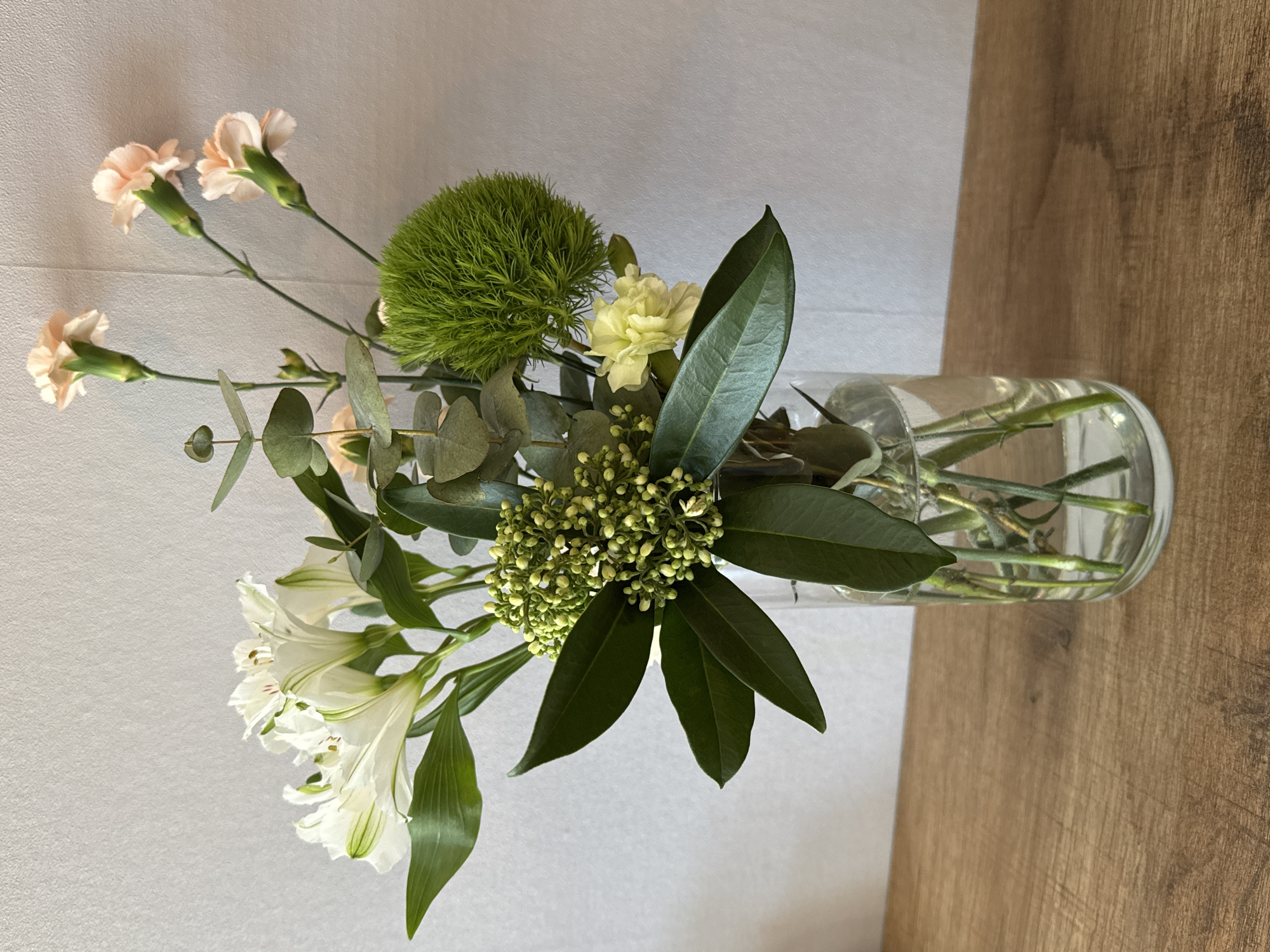 A top-down, still-life shot depicts a vase of flowers. The vase, a clear glass cylinder, contains water and the stems of the various flowers and foliage.
The arrangement includes:
- Several white alstroemeria flowers with green accents on the petals.
- A round, green ball-shaped flower resembling a dianthus green trick.
- Peach-colored carnations.
- Eucalyptus leaves, round and silvery-green.
- Dark green, glossy leaves.
- Small clusters of green buds.
The vase sits on a surface that is partially wooden (a light brown wood grain) and partially a plain white surface.
The location is likely indoors, but the specific location in the city of Chaikovsky, Russia cannot be determined from the visual information.